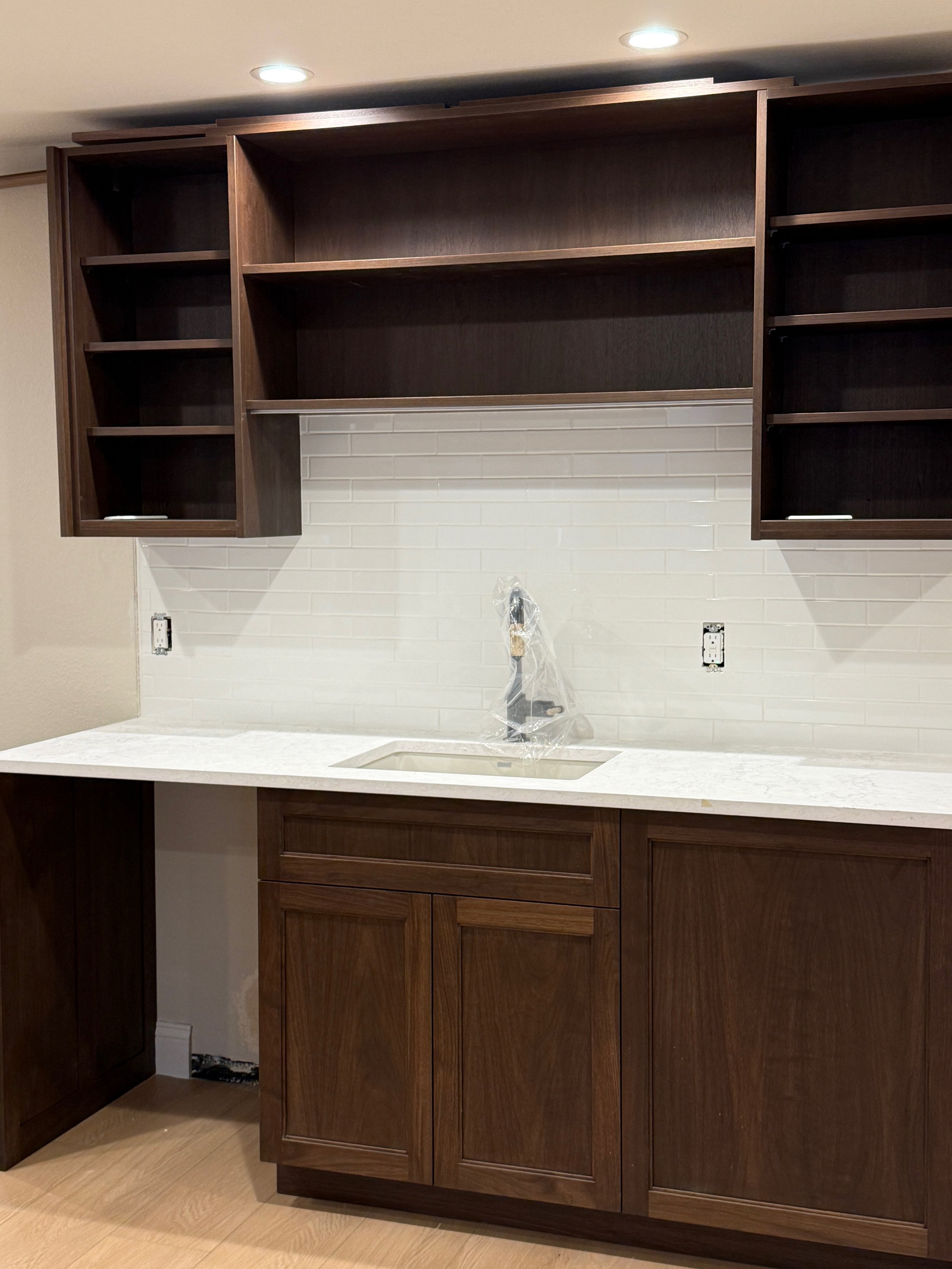 Kitchen with dark wood cabinets, white subway tile backsplash, and a light-colored countertop with an under-mount sink. Open shelves above the countertop and a central faucet wrapped in plastic. Two recessed ceiling lights overhead.