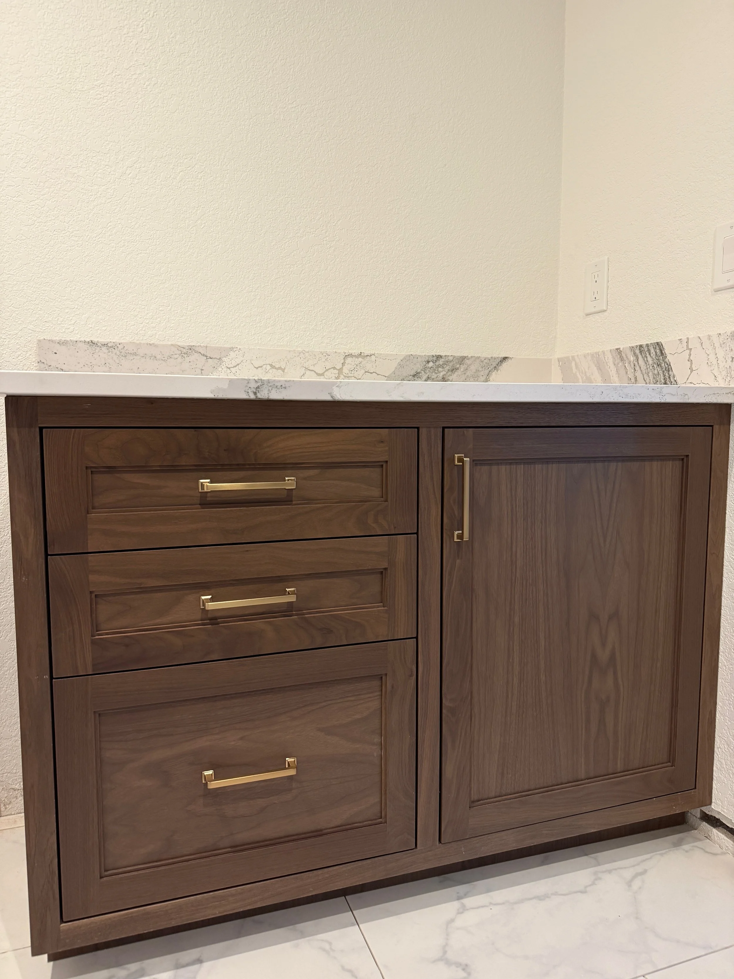 Wooden cabinet with gold handles and a marble countertop. The wall behind is beige with a textured finish, and there are electrical outlets on the right.