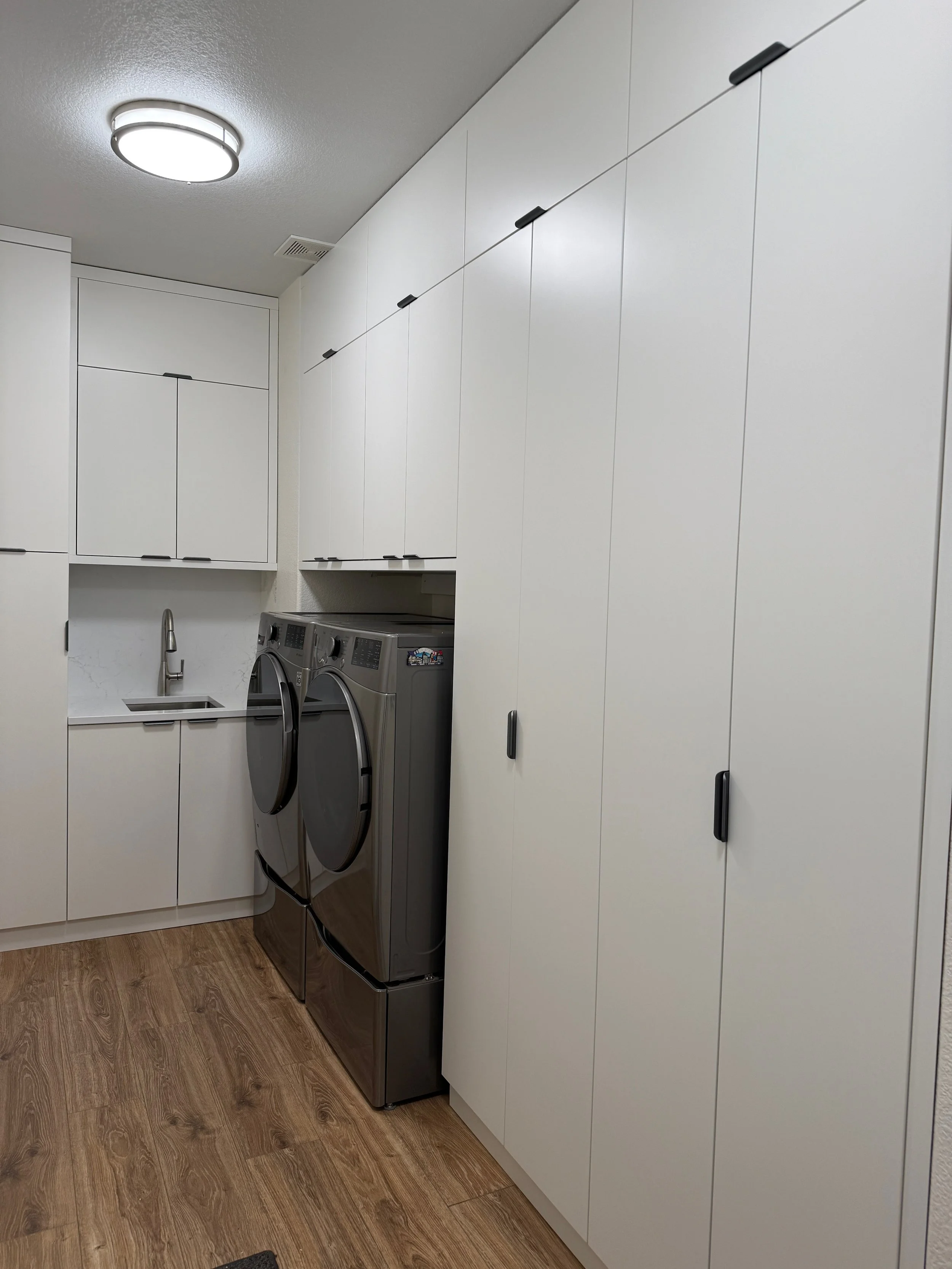 A laundry room with white cabinets, a stainless steel washer and dryer, a small sink with a faucet, and wooden flooring.