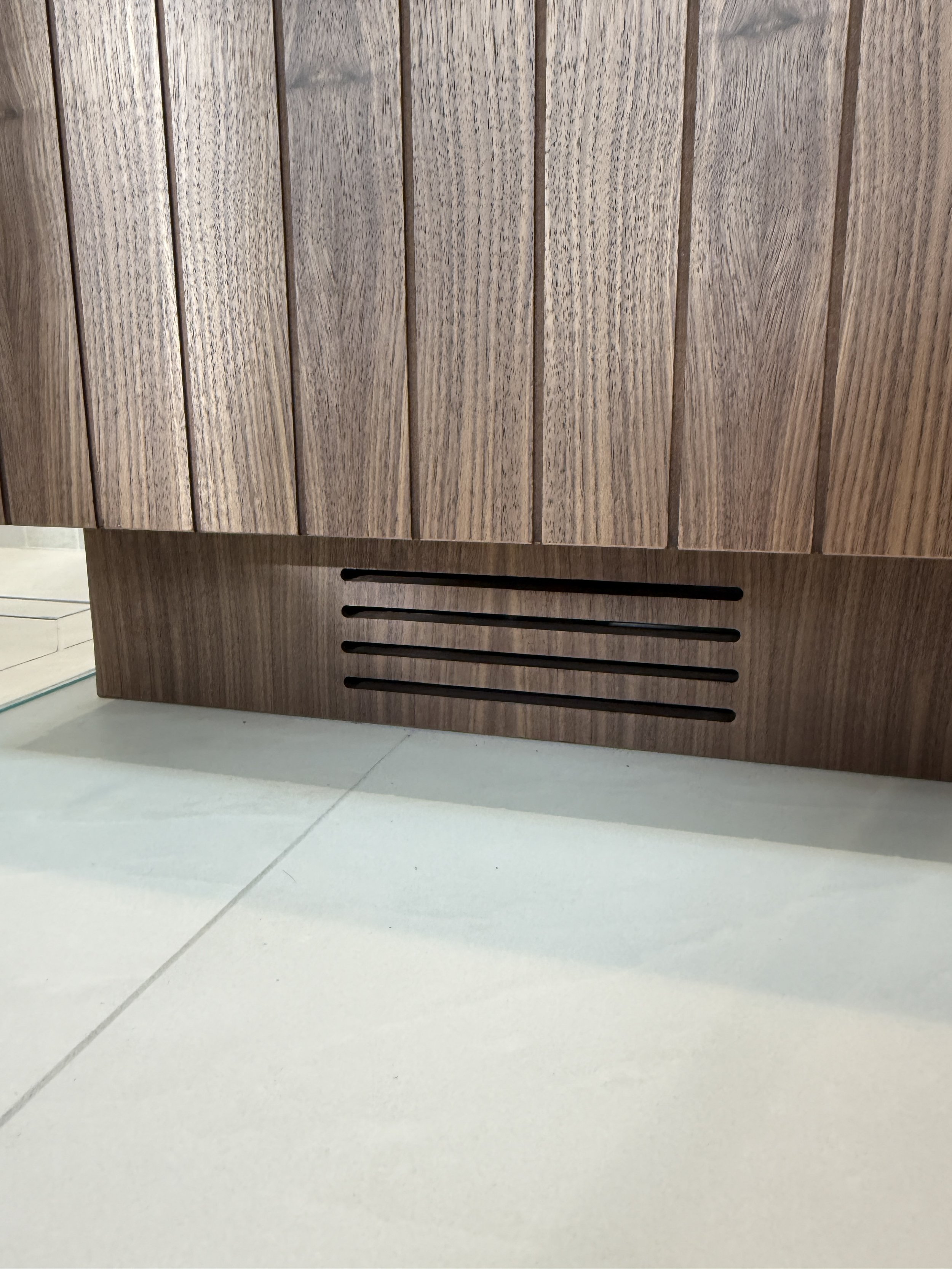 Close-up view of a wooden cabinet with vertical panels and horizontal vent slats near the floor, on a tiled floor.