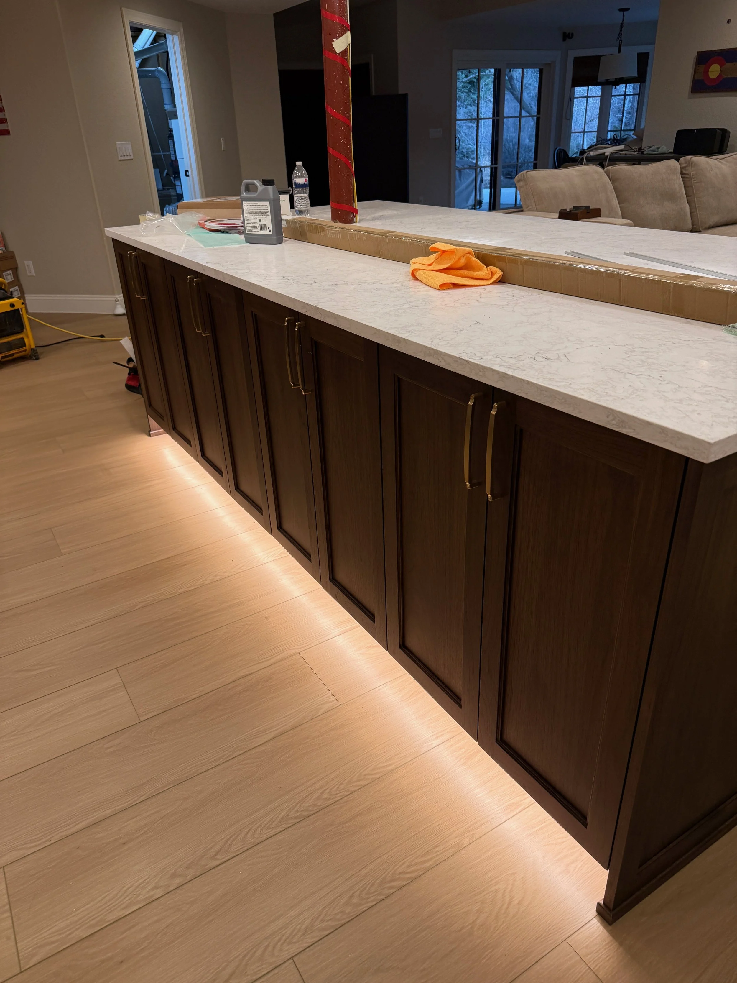 Kitchen island with a marble countertop and dark wood cabinets, with tools, a water bottle, and cleaning cloth on the counter. There is a red and white pillar at the back of the island, and the room has wooden flooring with LED strip lighting underne