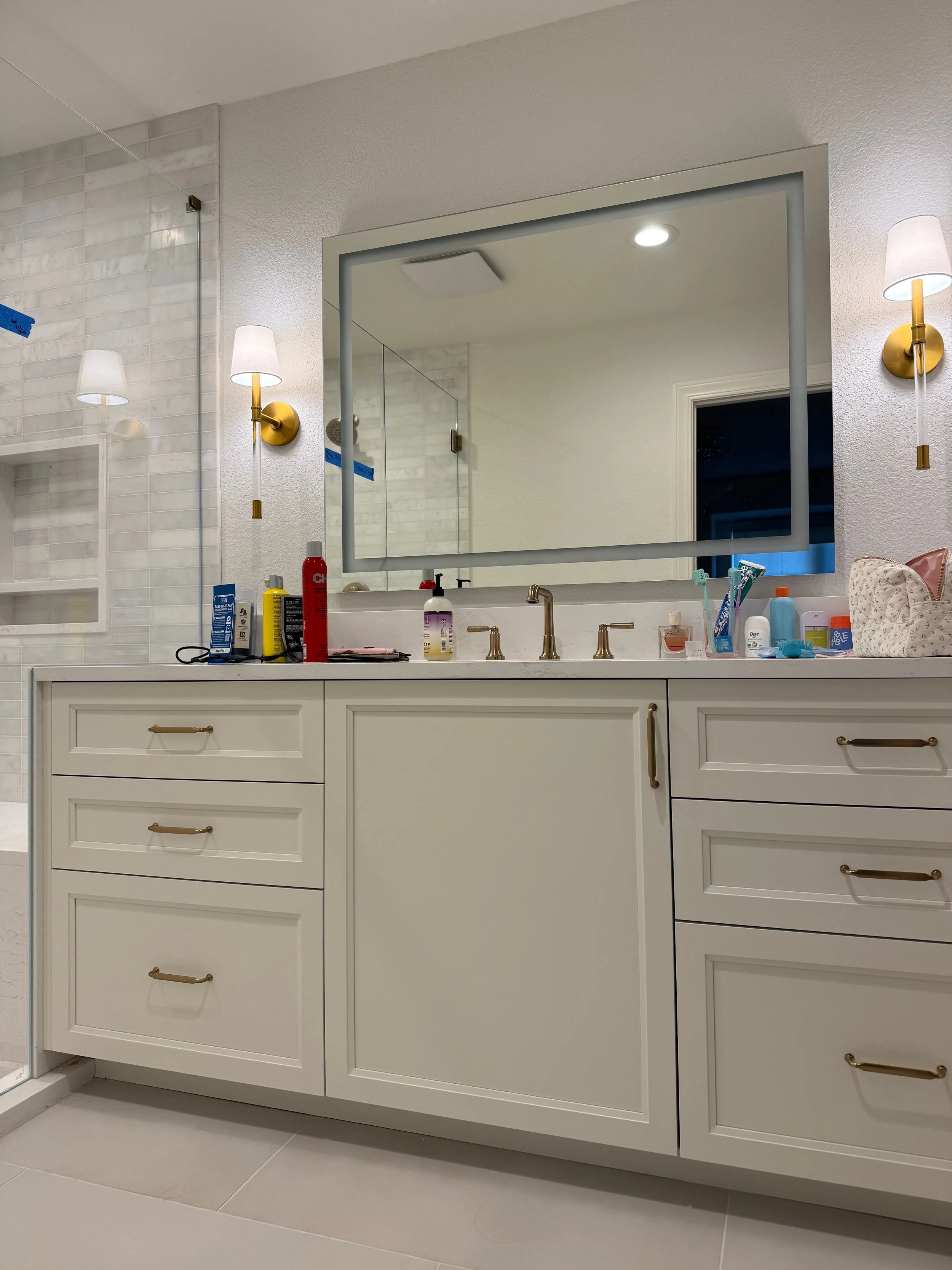 Bathroom vanity with various toiletries and a large wall mirror, illuminated by two gold and white wall sconces.