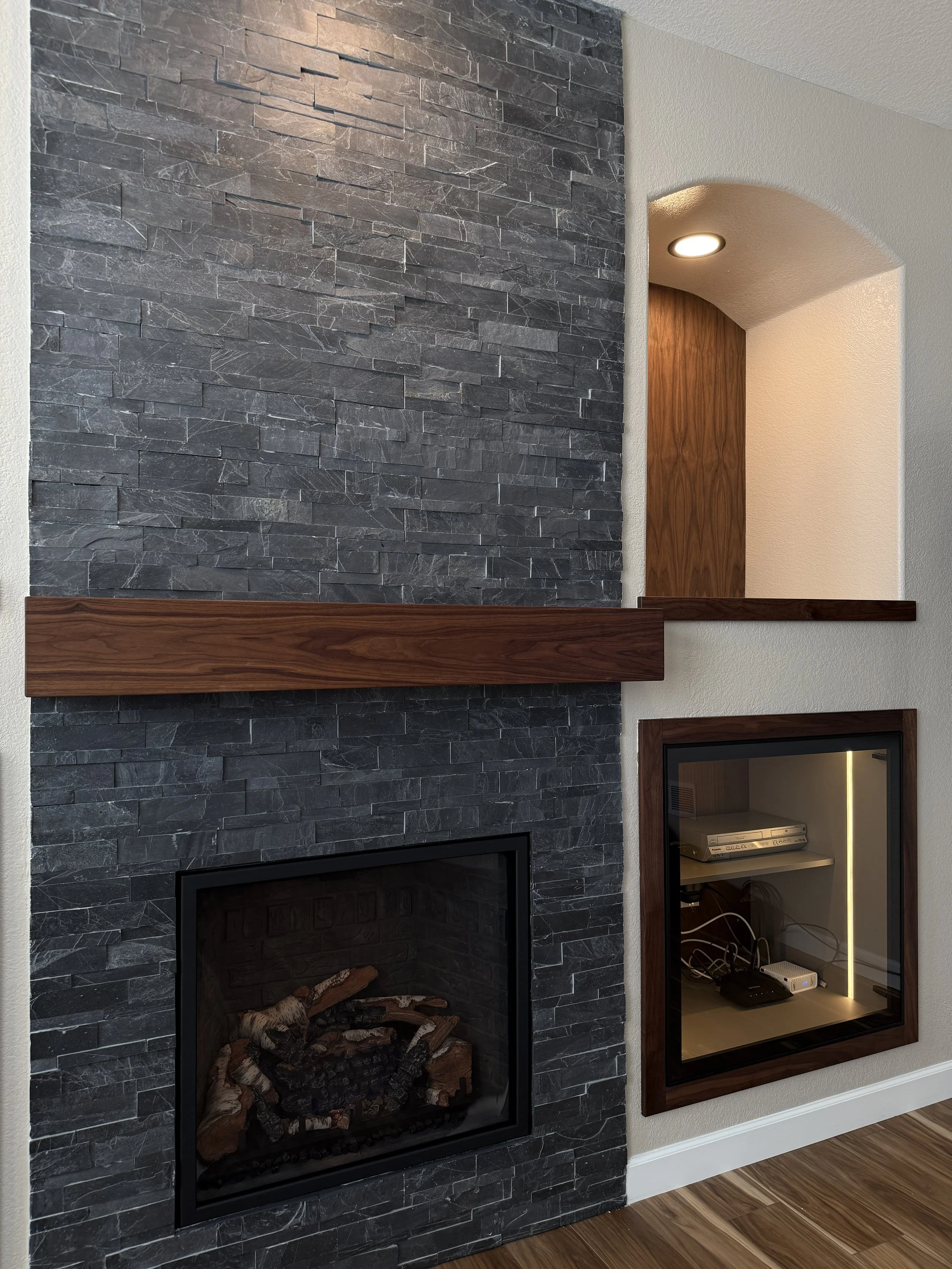 Interior of a living room featuring a modern black stone fireplace with a wooden mantle, adjacent to a built-in glass-covered electronic cabinet, under a small arched wall nook with a ceiling light.