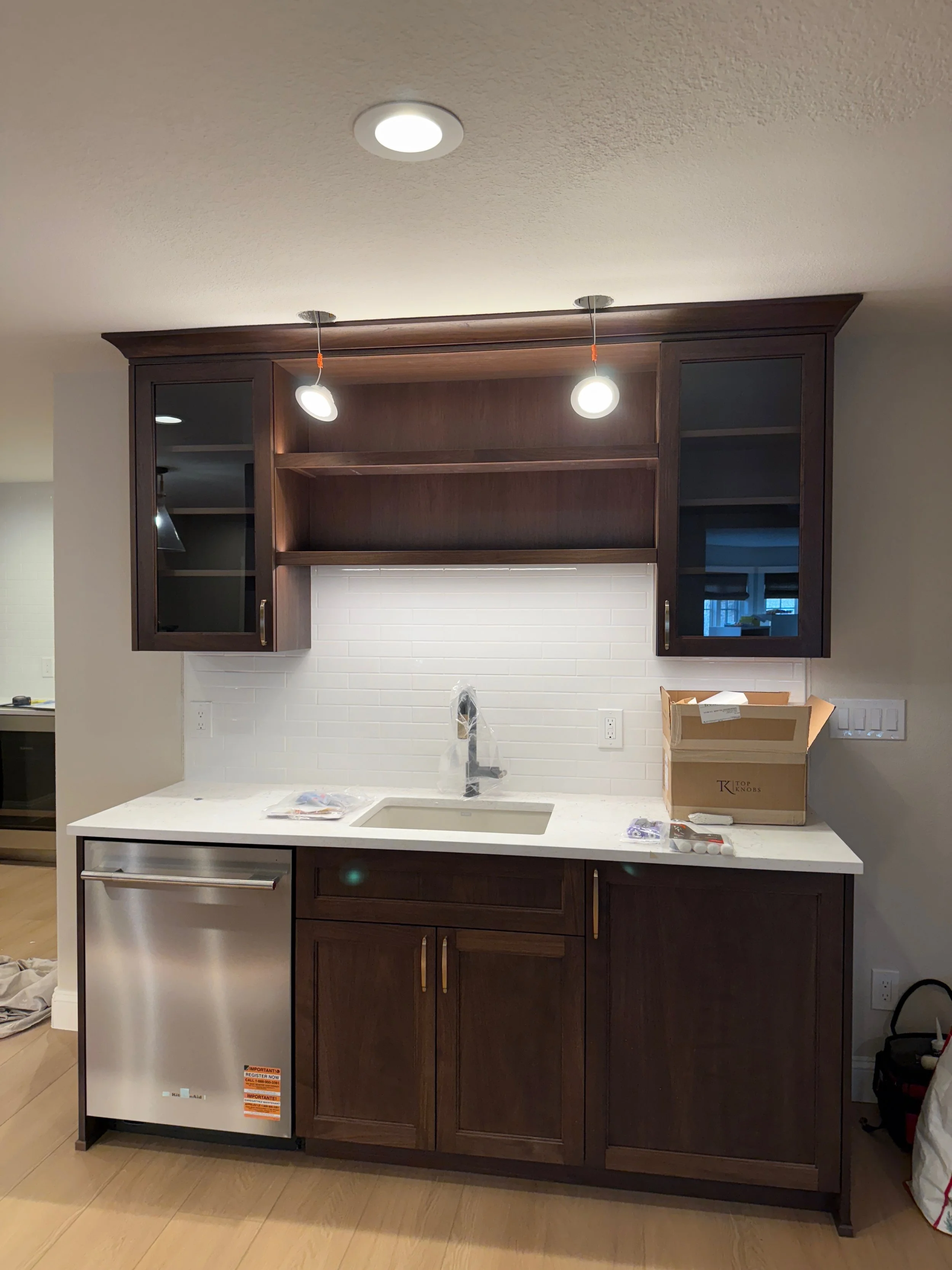Kitchen area with dark wood cabinets, a white countertop, a sink, and a small dishwasher, with open shelves and a white subway tile backsplash.