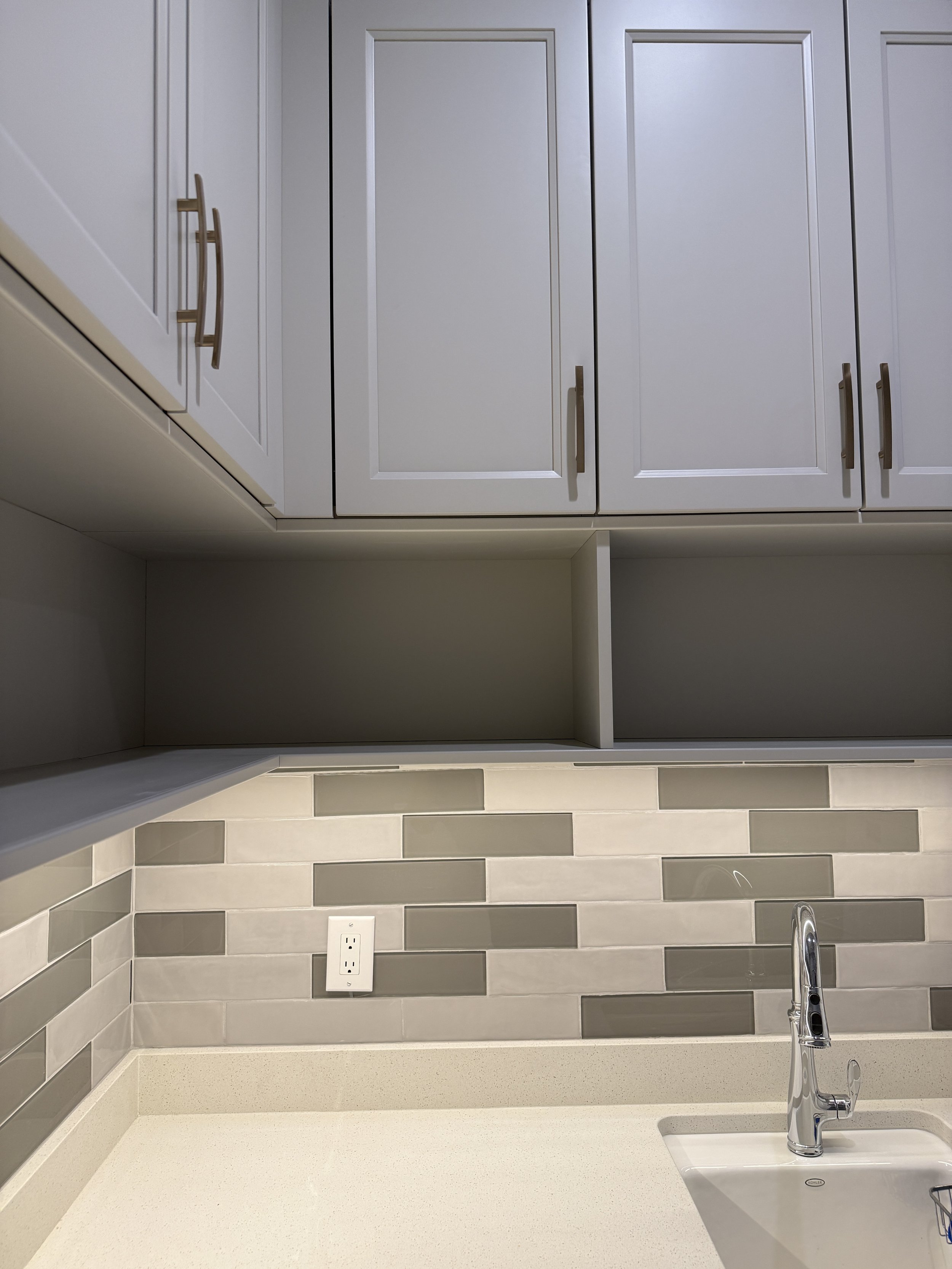 Kitchen countertop with a beige surface, backsplash with gray and beige bricks, an electrical outlet, and a stainless steel faucet above a sink. Upper cabinets are white with gold handles.