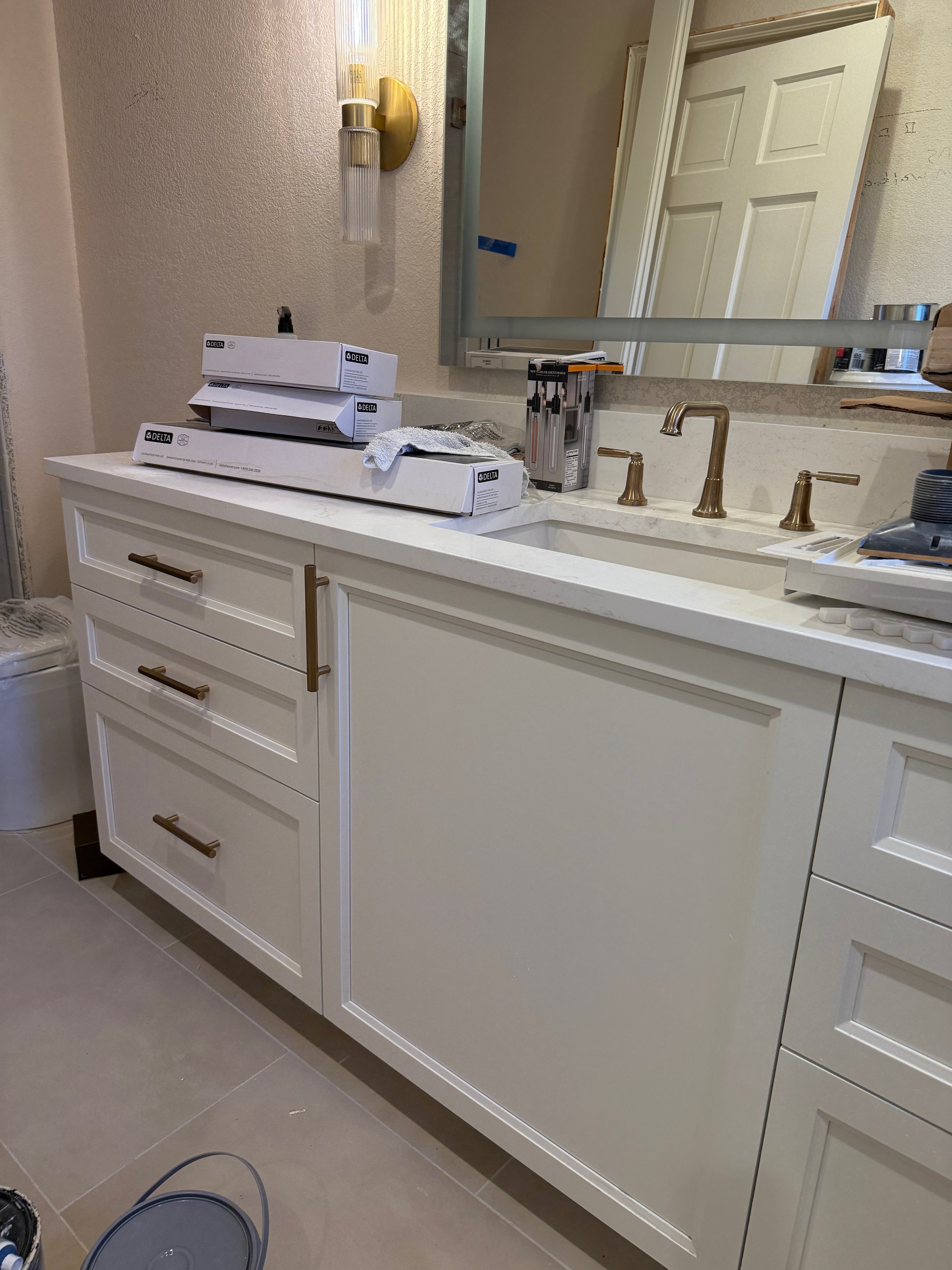 Bathroom vanity with three boxes of Delta faucet parts, cleaning cloth, and tools on the counter, gold fixtures, and a large mirror above.