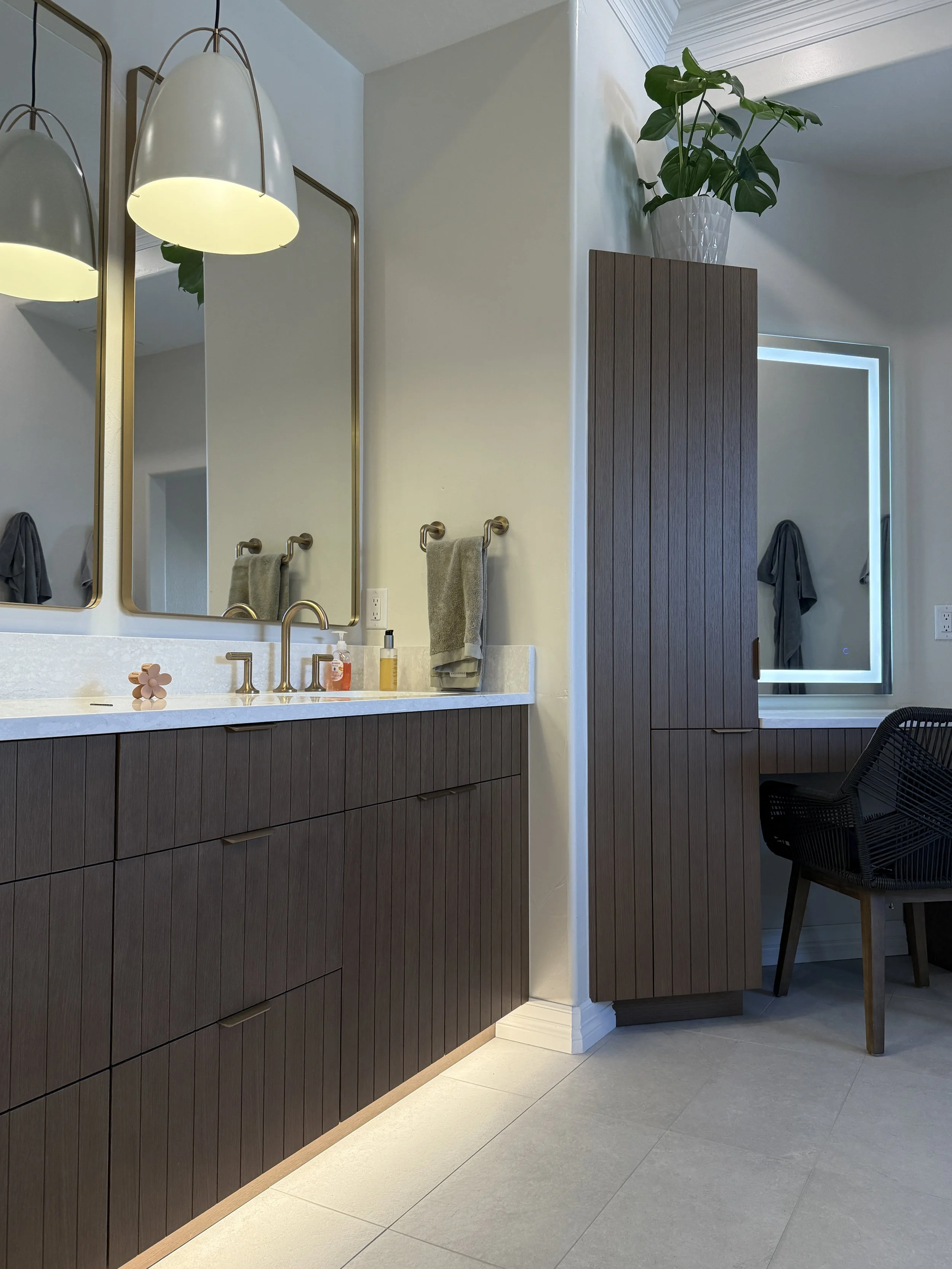 Modern bathroom with dark wood cabinets, large mirrors, and a white countertop with a sink. Towel hooks with gray towels, a potted plant on a tall wooden cabinet, and a black chair near a backlit mirror.