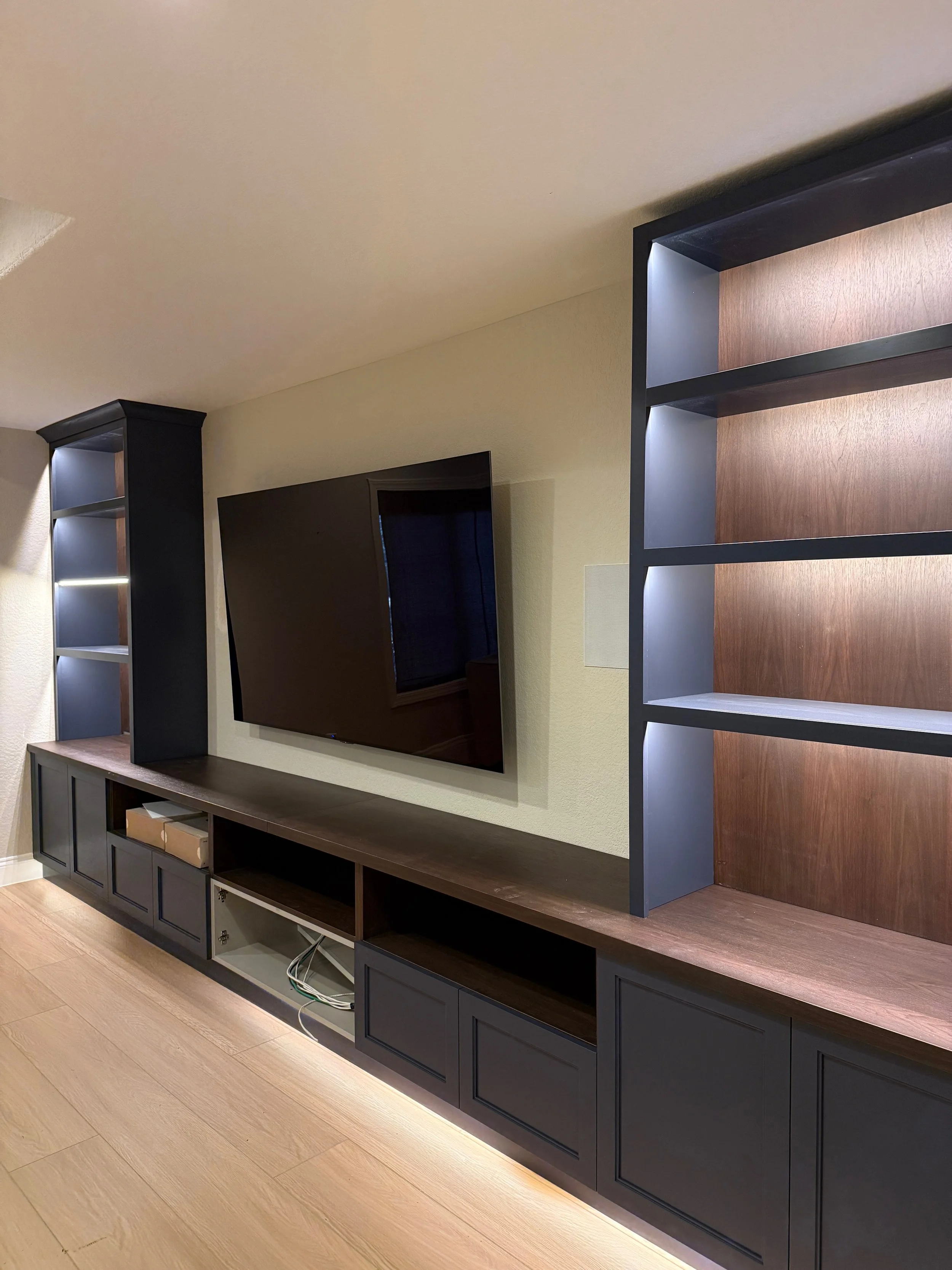 Modern TV entertainment center with black cabinetry, featuring open shelves and closed cabinets, a wall-mounted TV, and decorative lighting, in a room with light-colored wooden flooring.