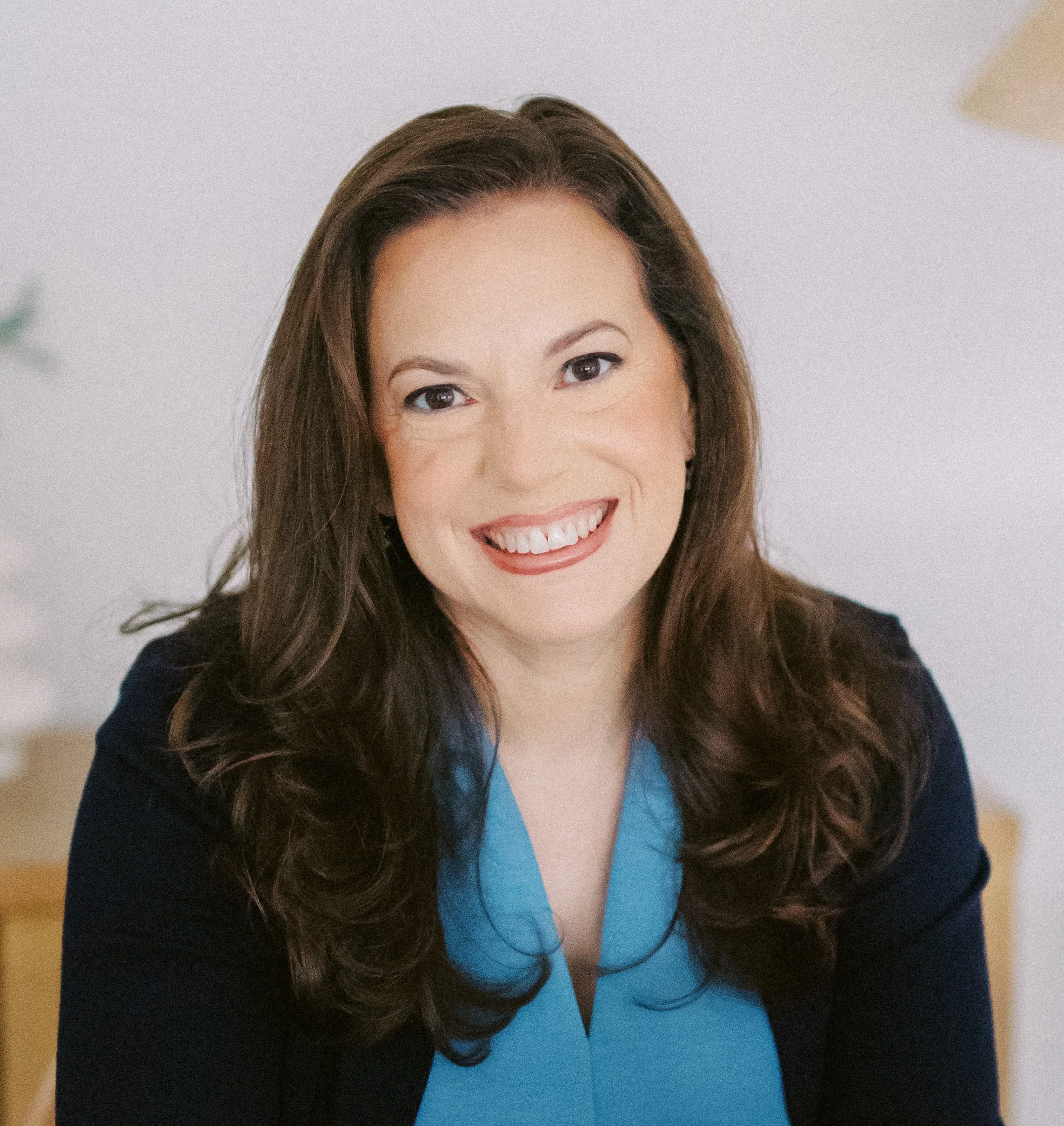 Portrait of a smiling woman with long dark hair, wearing a blue blouse and black blazer, indoors.