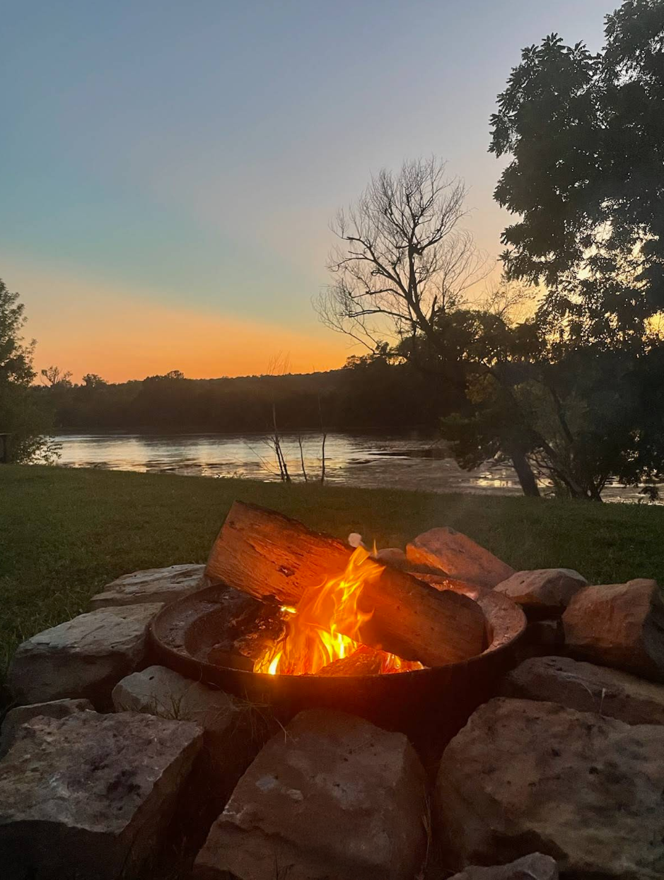 A campfire surrounded by rocks near a river at sunset with trees on the shoreline and a partly cloudy sky.