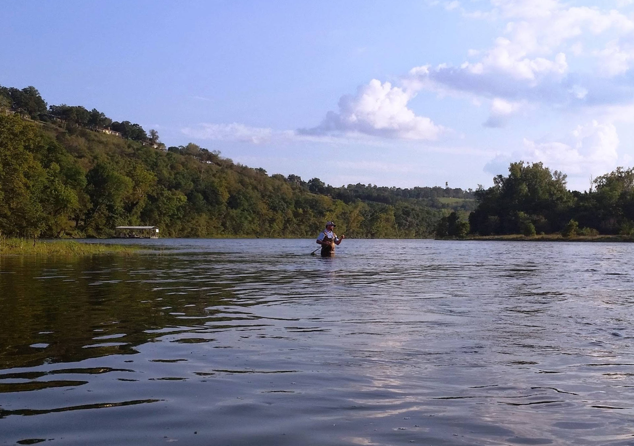 A person fishing in a wide river with green hills and trees on the banks under a partly cloudy sky.