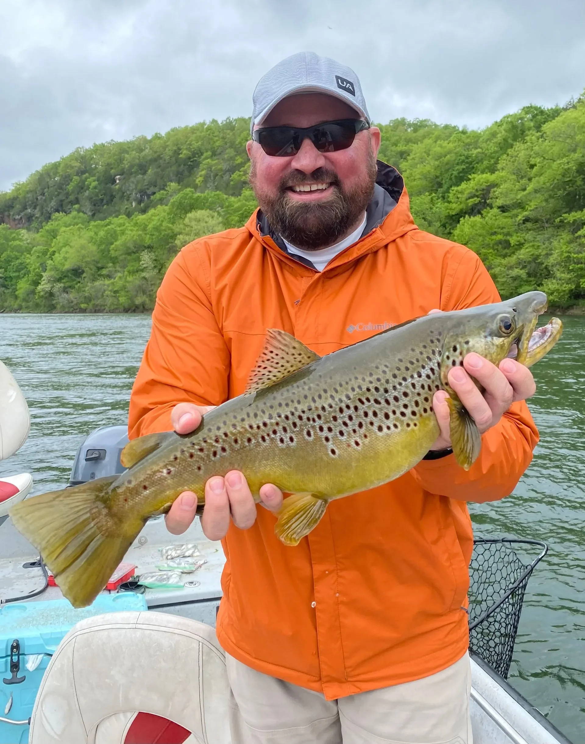 A man wearing sunglasses, a gray cap, and an orange jacket holding a large brown trout fish on a boat with water and green trees in the background.