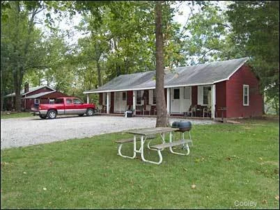 A red house with a gray roof in a wooded area. In front, there is a gravel driveway with a red pickup truck parked on it. A picnic table with a grill is on the lawn in front of the house.