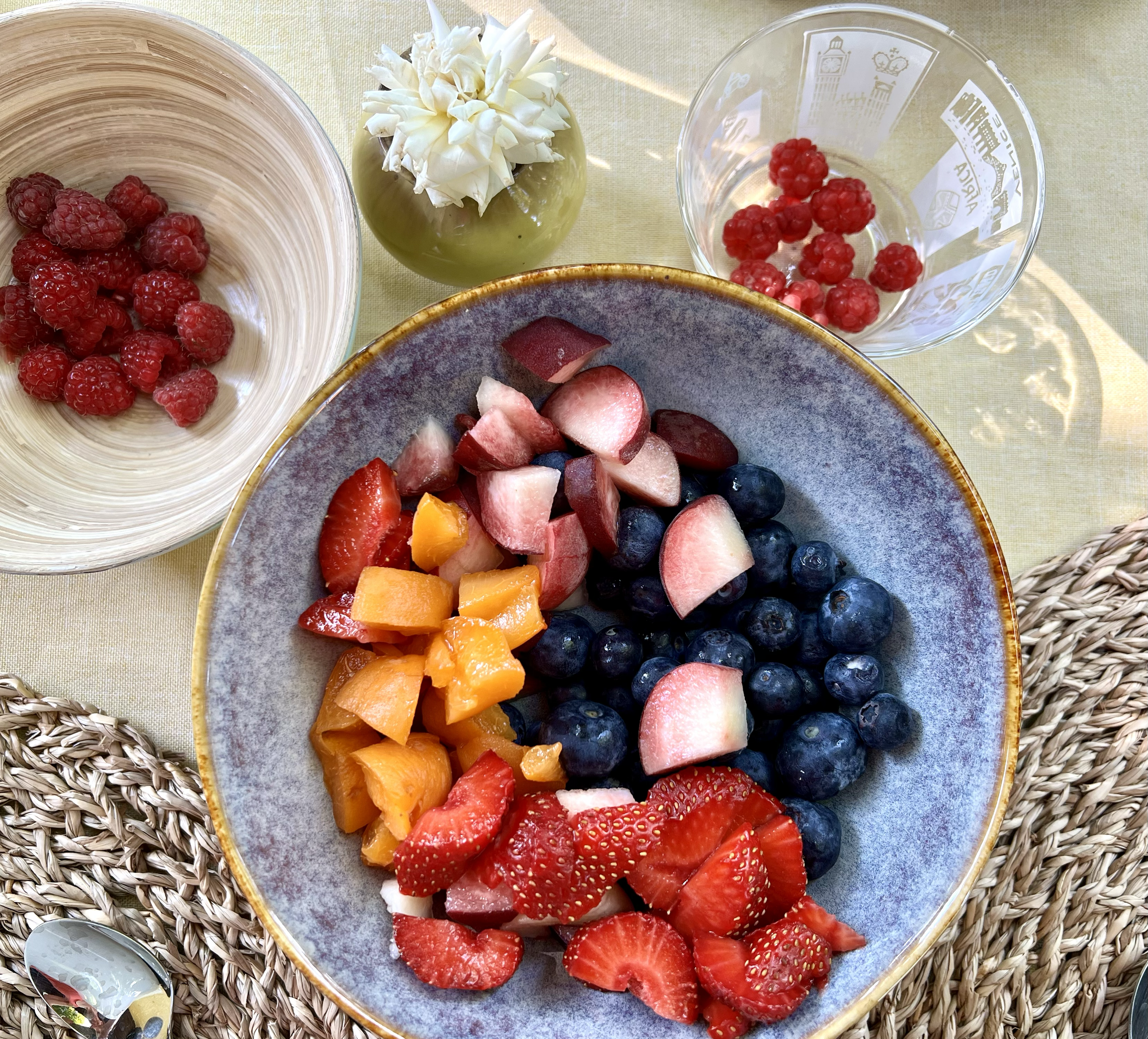 Eine Schale mit verschiedenen frischen Früchten wie Erdbeeren, Heidelbeeren, Wassermelone, Honigmelone, Pfirsich und Papaya. Daneben eine weitere Schale mit Himbeeren und Rote-Bete-Stücken, eine Vase mit weißen Blumen, und ein Löffel auf einer Decke 