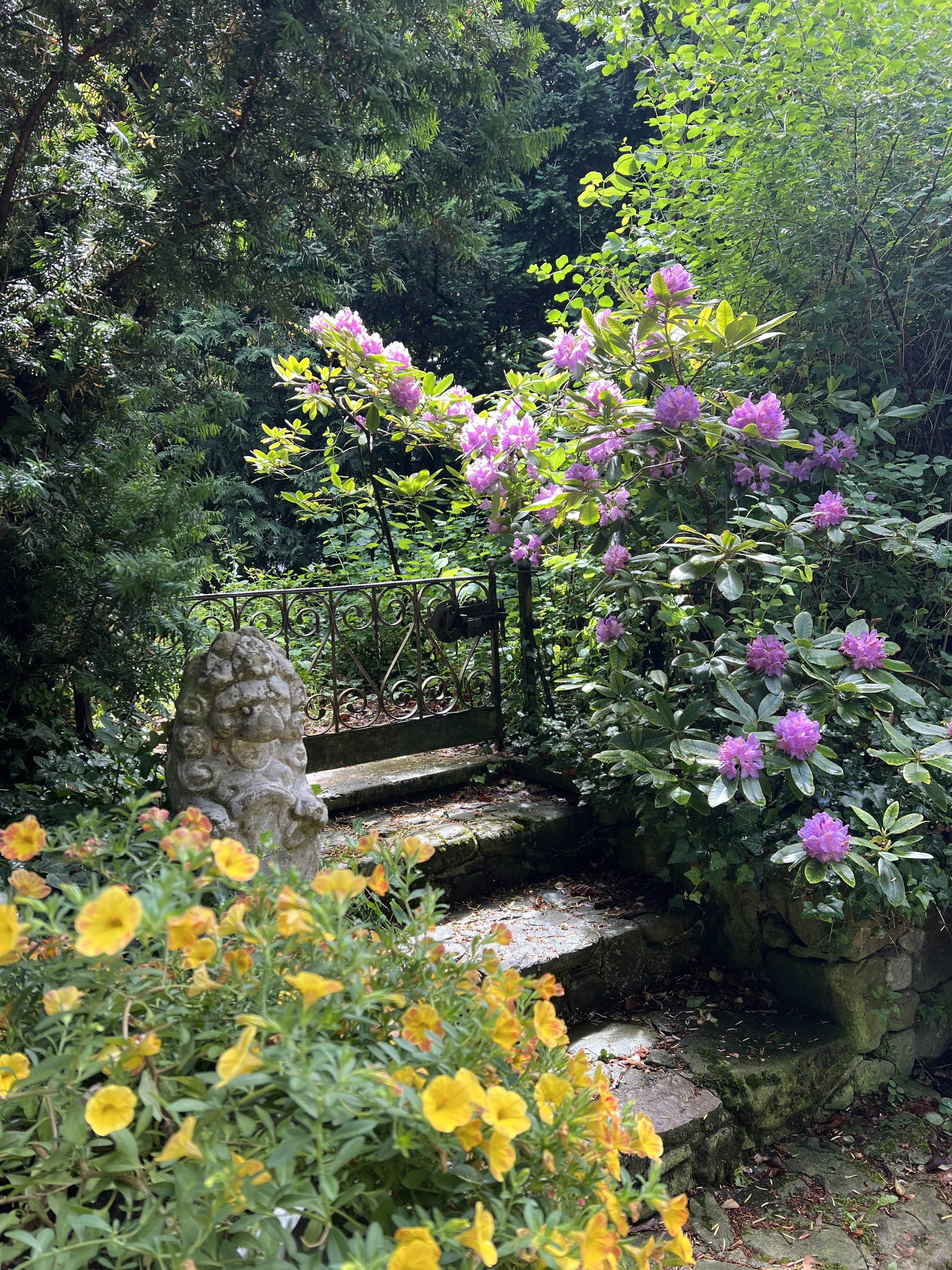 Ein Garten mit bunter Blumen, einem kleinen Wasserlauf und einer Hüttenbrücke, umgeben von dichter Vegetation.