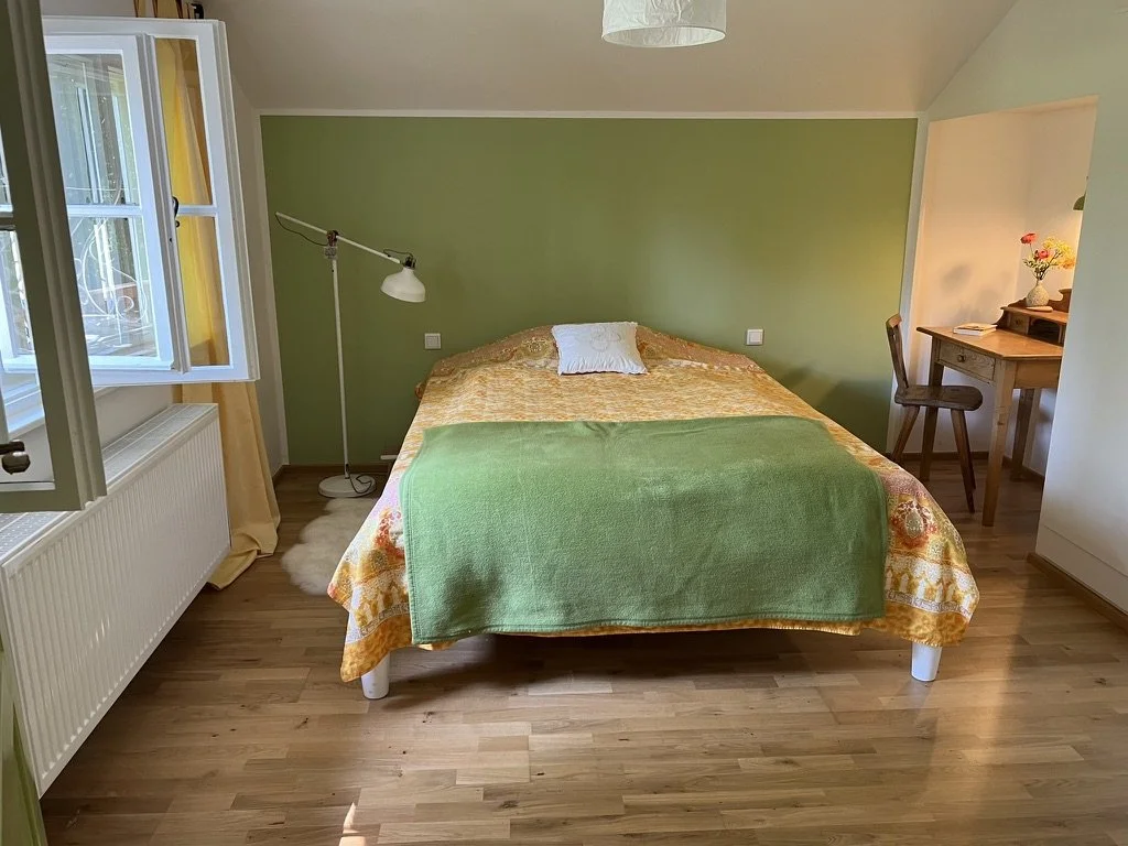 Ein Schlafzimmer mit grünem Wand, einem Doppelbett mit orangefarbenem Bettzeug und grünem Überwurf, einer Stehlampe, einem kleinen Schreibtisch mit Stuhl und einer offenen Fensterscheibe mit gelben Vorhängen.