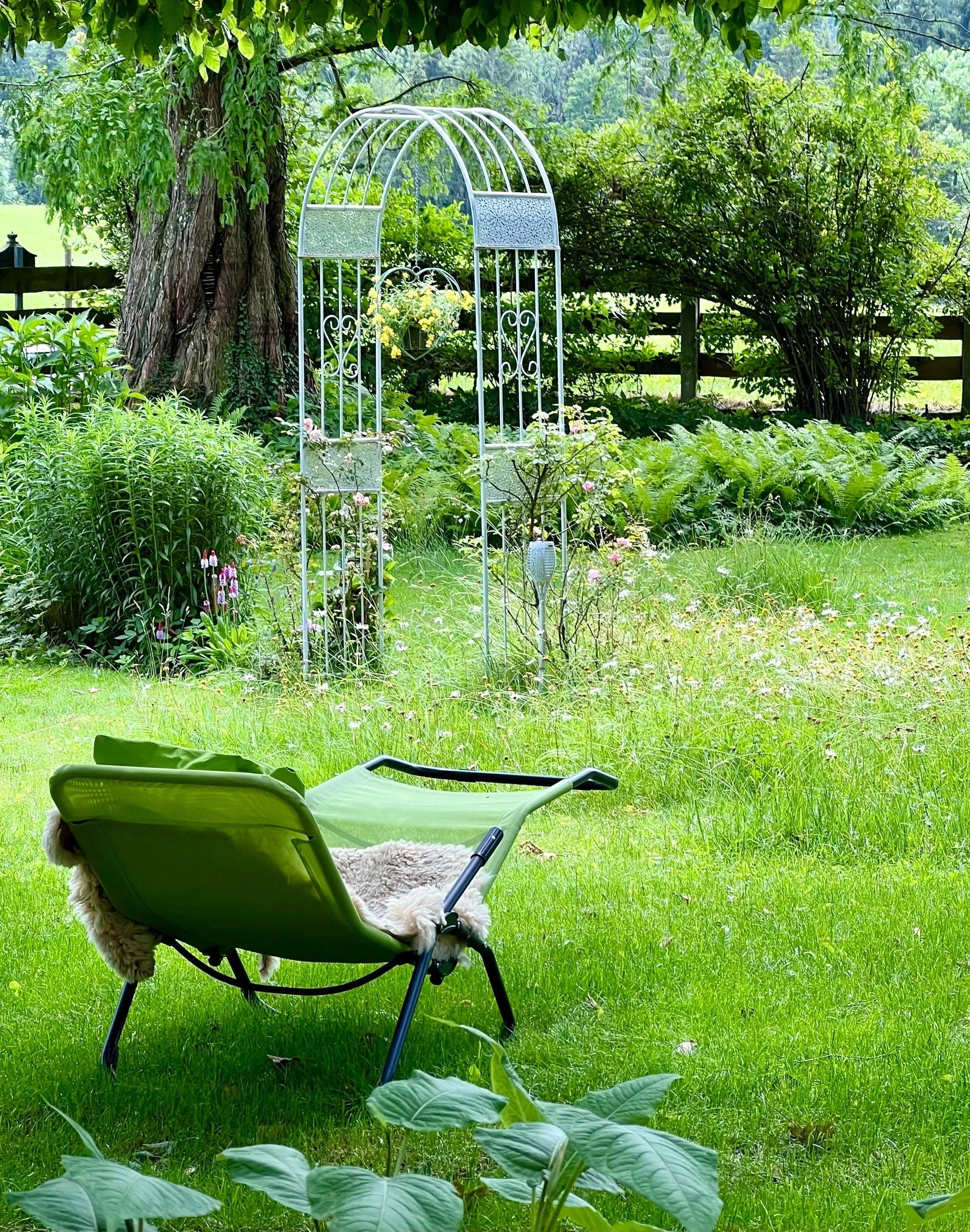 Garten mit liegendem grünen Liegestuhl, verzierter weißer Gartenlaube, Pflanzen und Bäumen im Hintergrund.