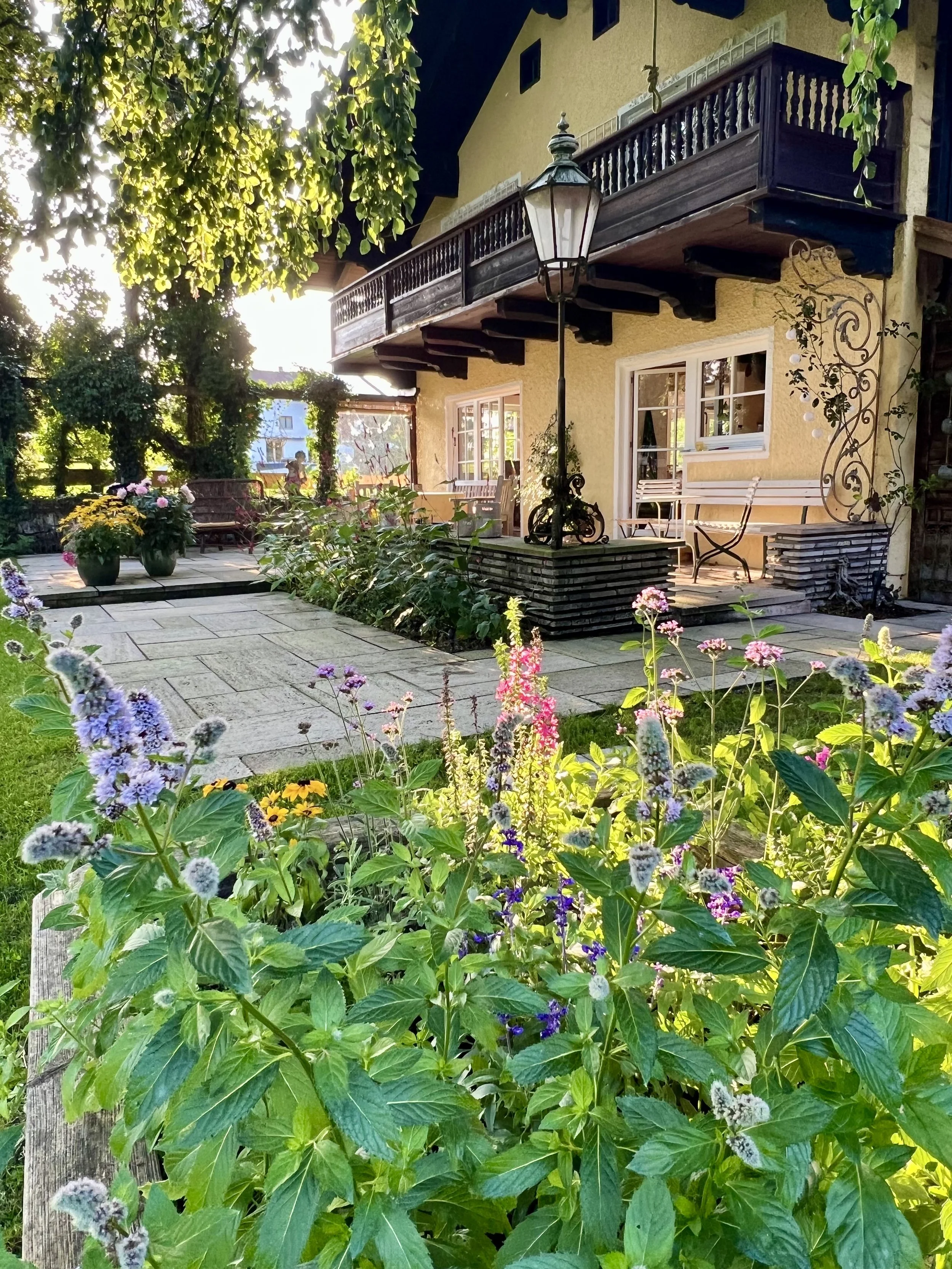 Blumen im Garten vor einem Haus mit Terrasse, Balkon und einer Laterne.