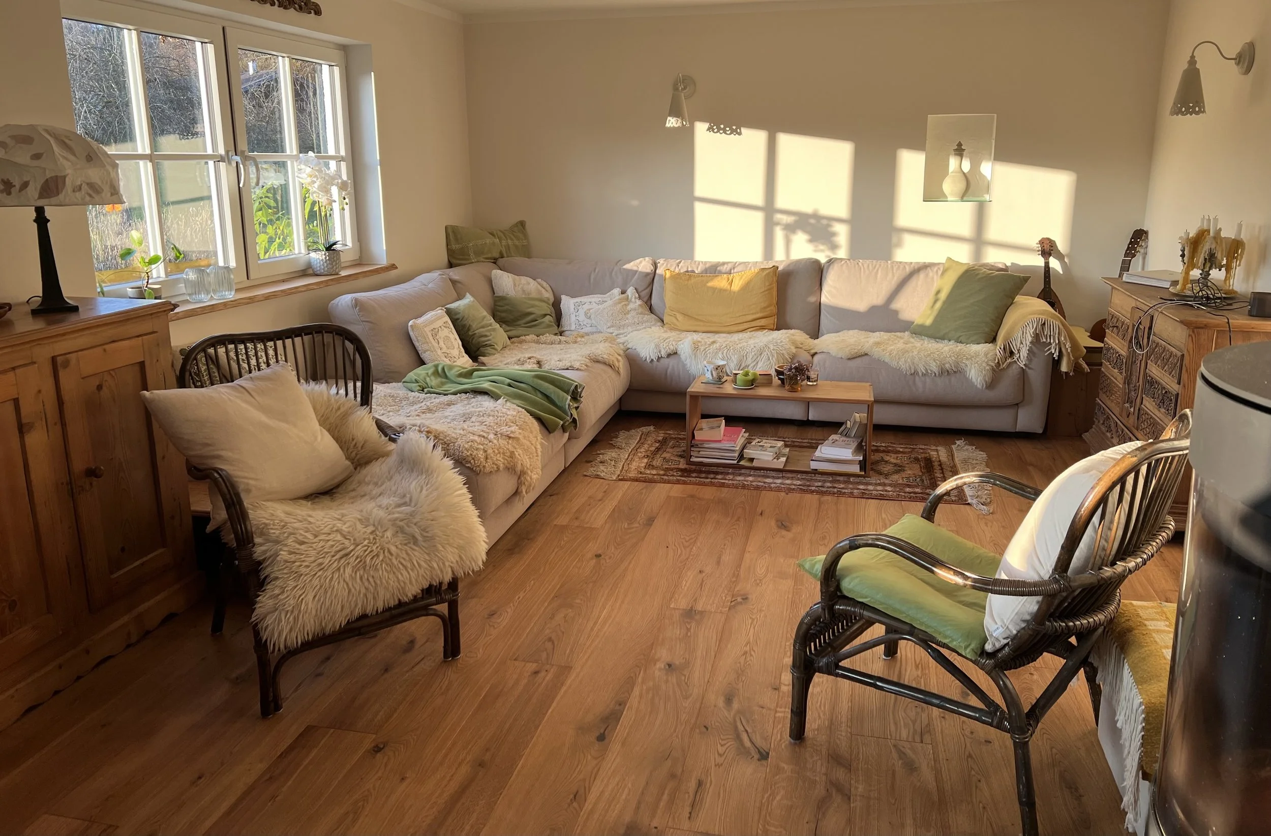 Gemütliches Wohnzimmer mit einem großen hellen Sofa, Kissen, einem Couchtisch mit Büchern und Dekoration sowie mehreren Sesseln und Pflanzen bei Sonnenlicht.