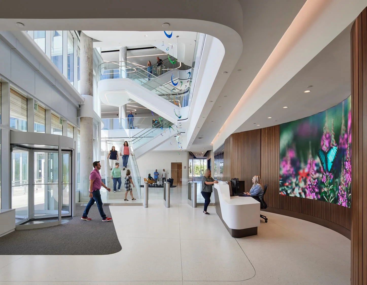 Interior of a modern building lobby with a reception desk, a large digital butterfly screen, a curved staircase, and multiple people walking and interacting.