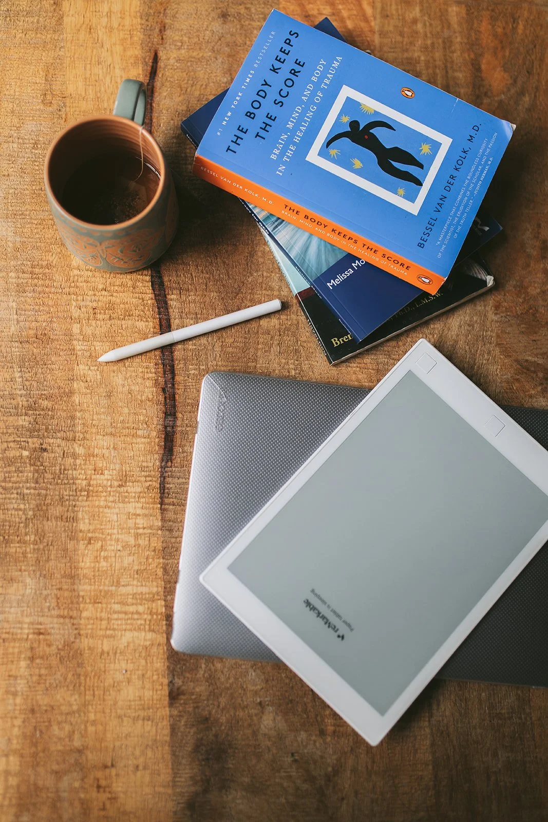 Flat lay of clinical books including The Body Keeps the Score, a reMarkable tablet, a stylus, and a ceramic mug on a wood table