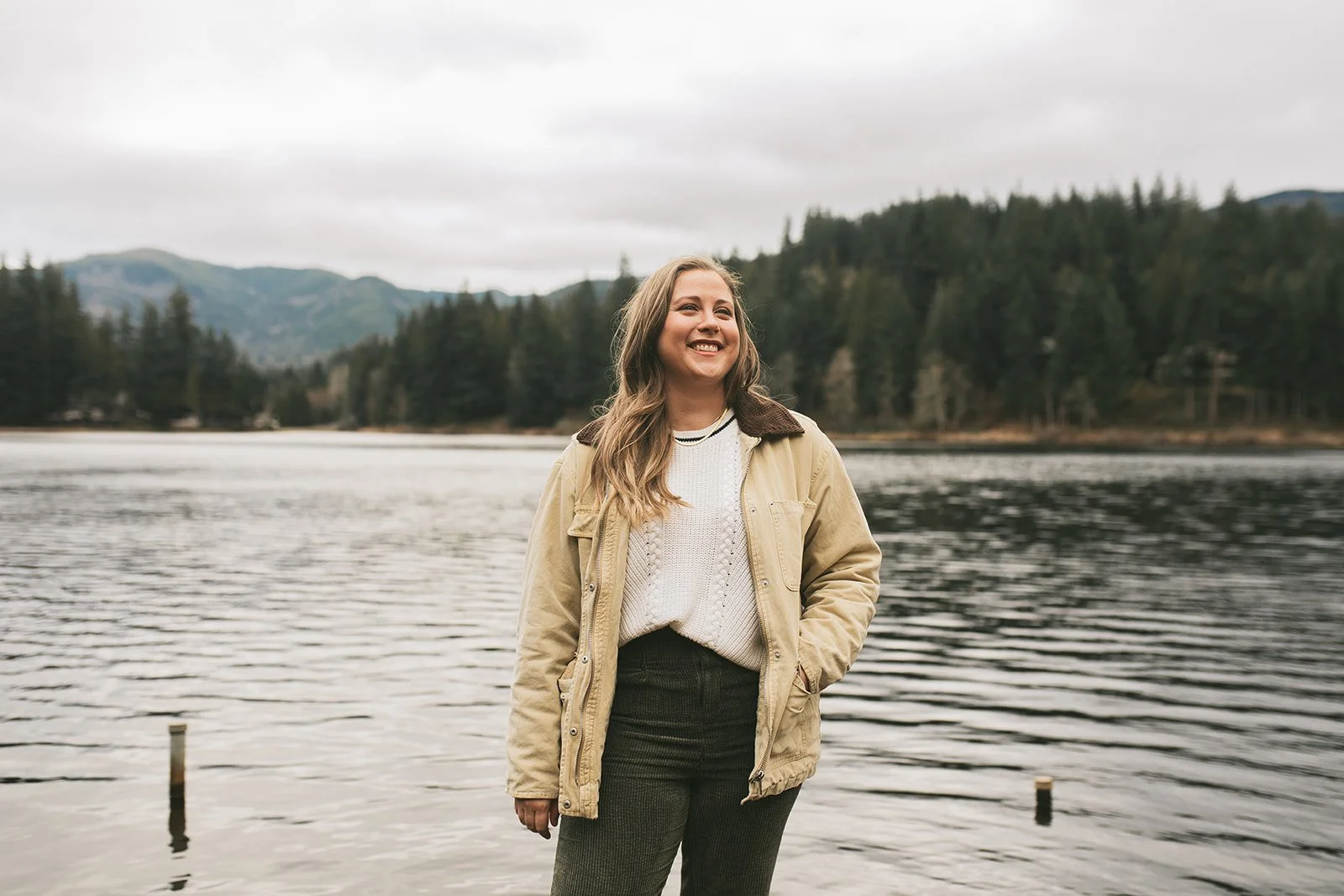 Meredith Garreau, founder of Wanderwell Counseling, smiling and looking up while standing at the edge of a lake with mountains and evergreen trees behind her