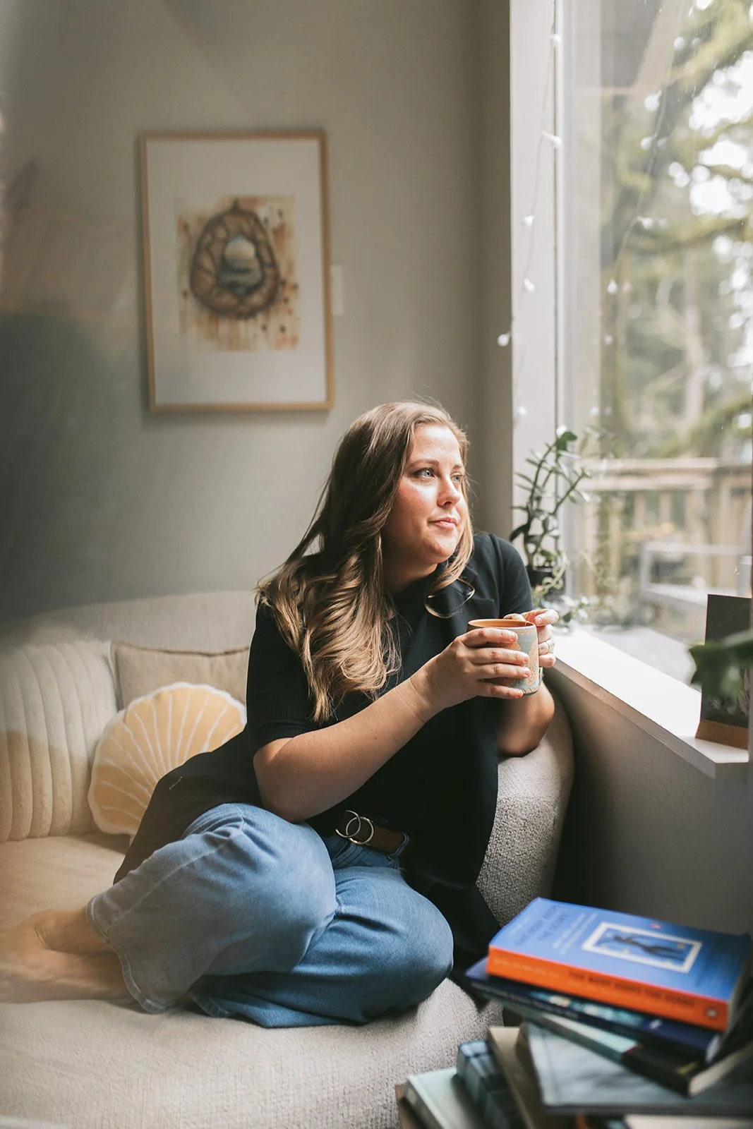 Meredith Garreau, founder of Wanderwell Counseling, sitting in a window seat holding a mug and gazing thoughtfully outside, with clinical books and a plant nearby