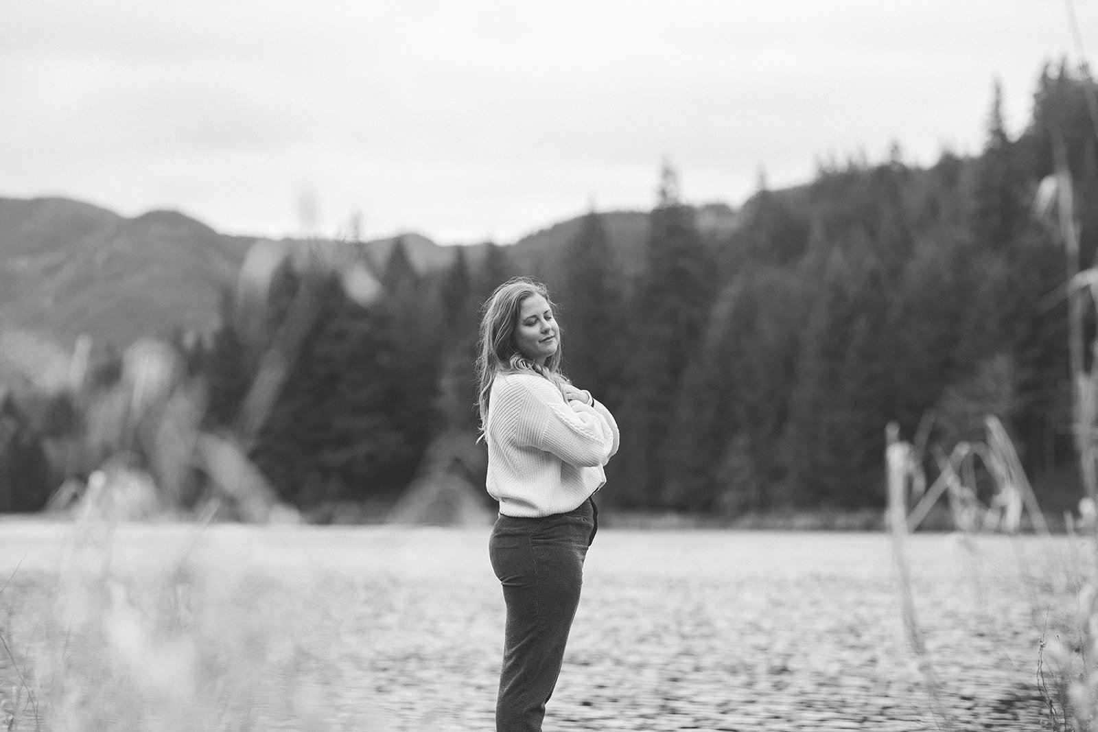 Black and white photo of Meredith Garreau standing at a lakeside with arms wrapped around herself, eyes closed, surrounded by mountains and evergreen trees