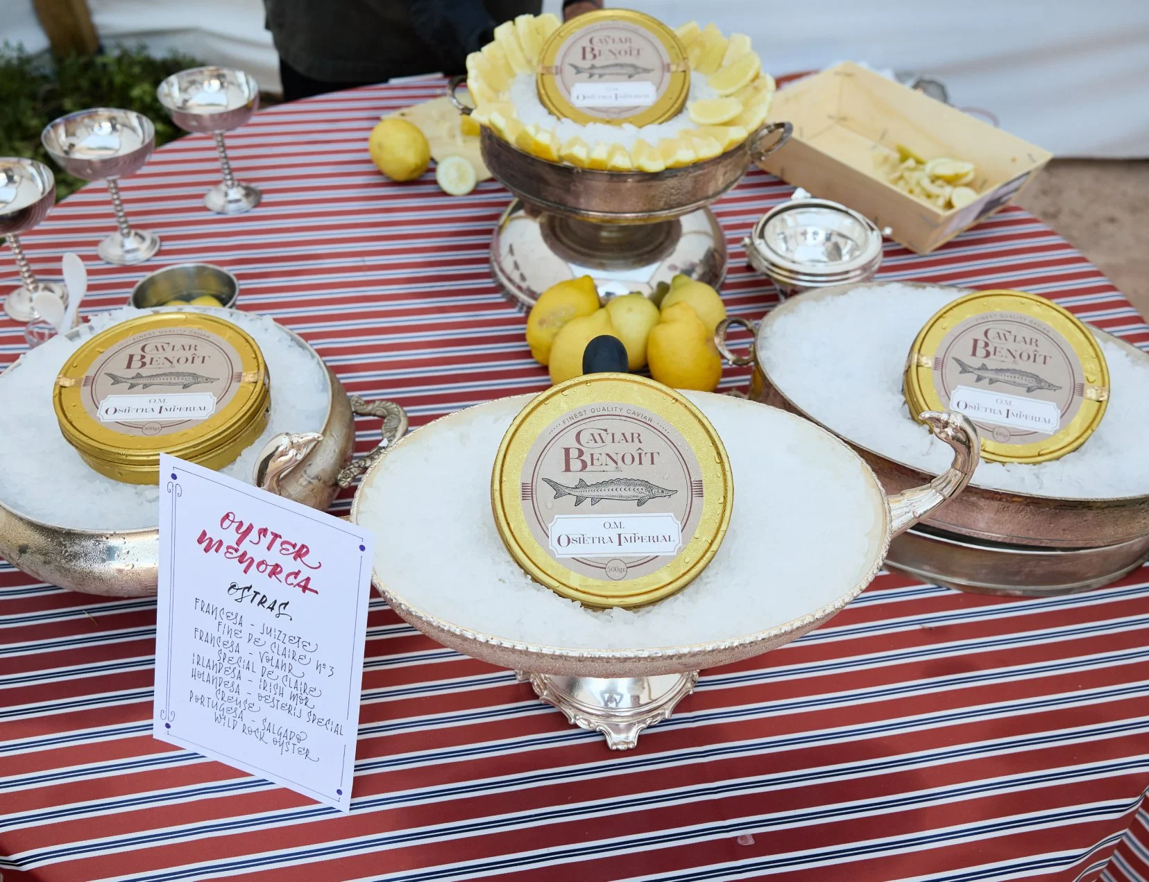 Several tins labelled 'Caviar Benoît' displayed on a decorative tray with a sign listing various types of oyster, on a red and white striped tablecloth with lemons and small cups in the background.