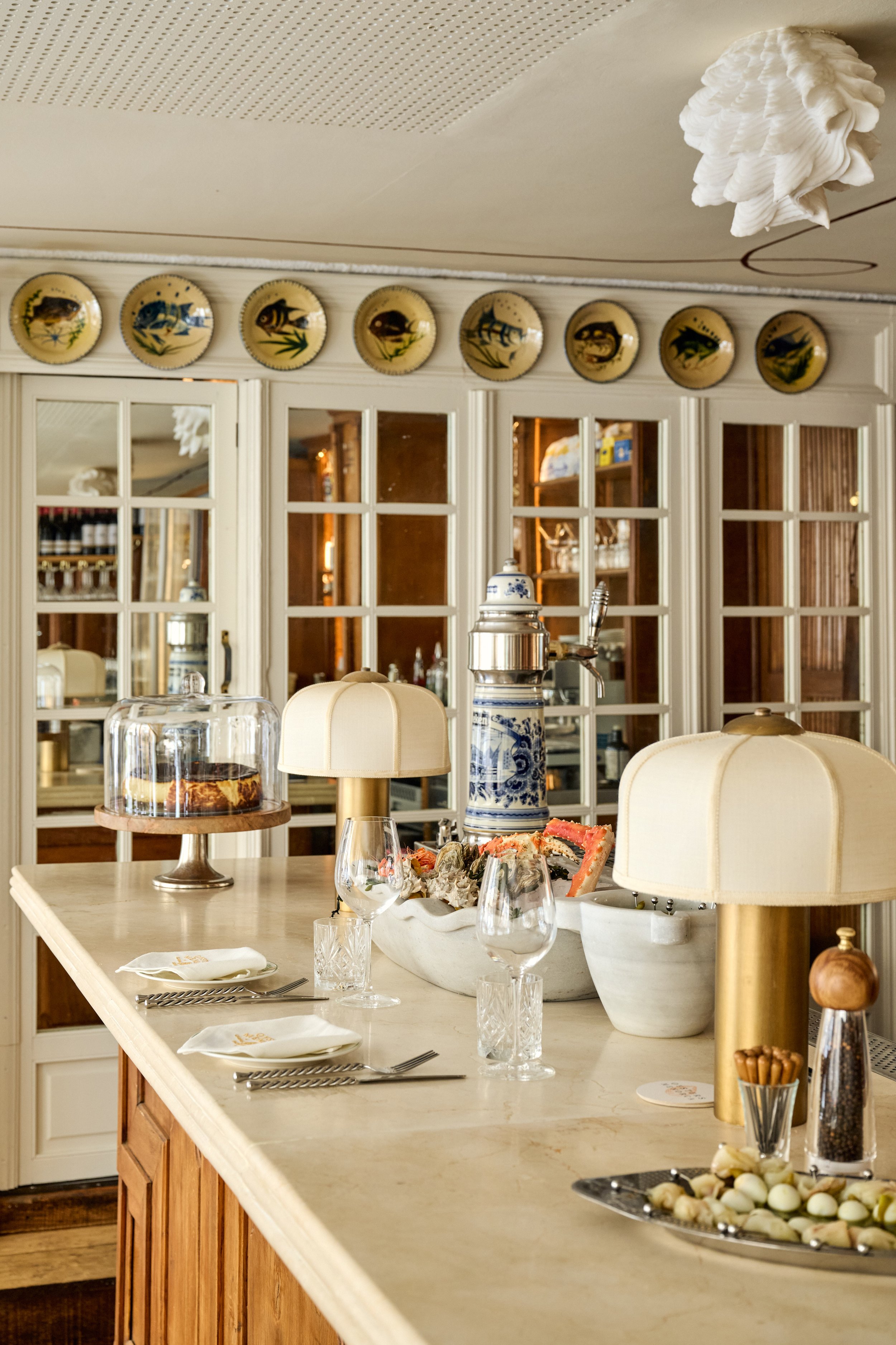 A dining room with a marble countertop, glassware, and two beige lamps. The background features a wooden cabinet with glass doors and a decorative wall with plates displaying fish. There is a cake under a glass dome, a ceramic coffee percolator, and 