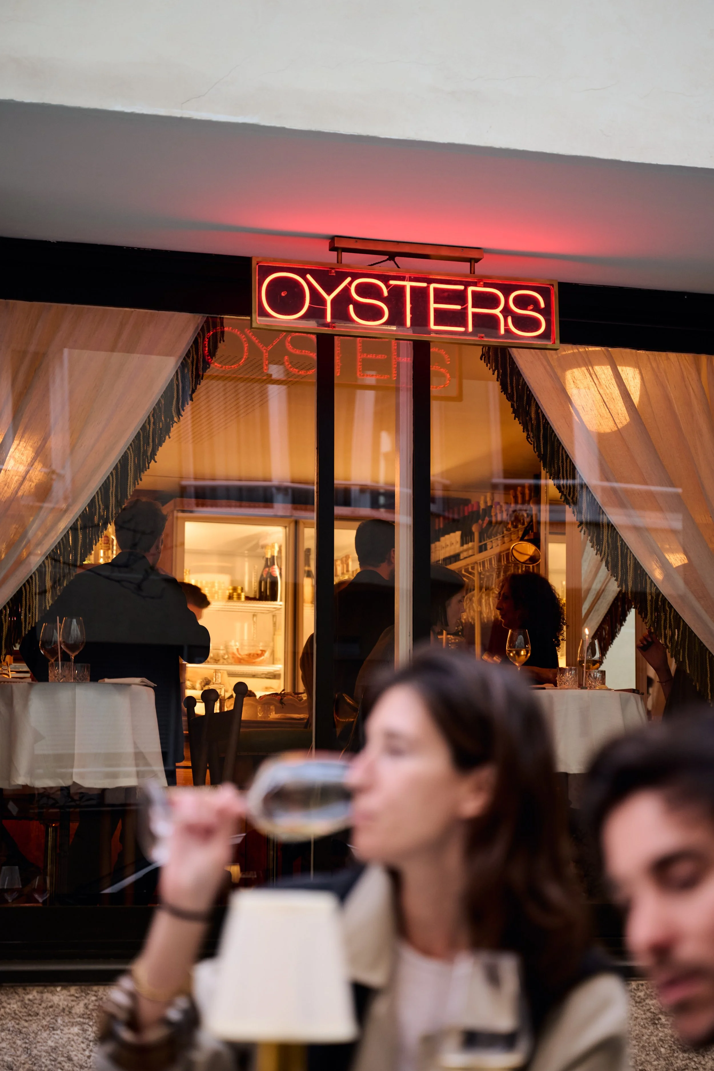 A neon sign that reads 'OYSTERS' hanging outside a restaurant, which has a view of the interior with people dining and wine glasses on the tables.
