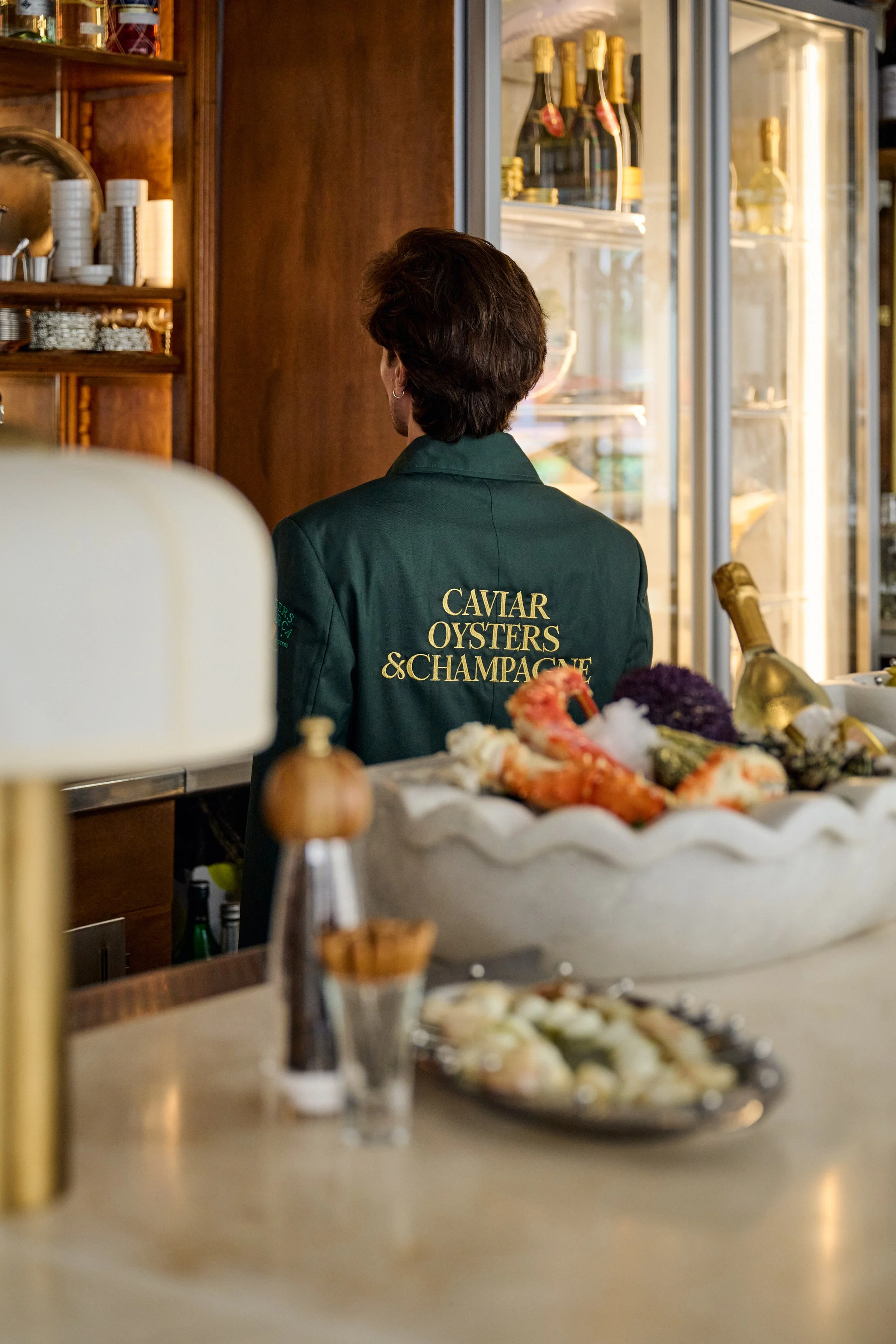 Woman working behind the bar at a seafood restaurant, wearing a jacket with 'CAVIAR OYSTERS & CHAMPAGNE' on the back, with a display case of champagne bottles and a bowl of seafood in the foreground.