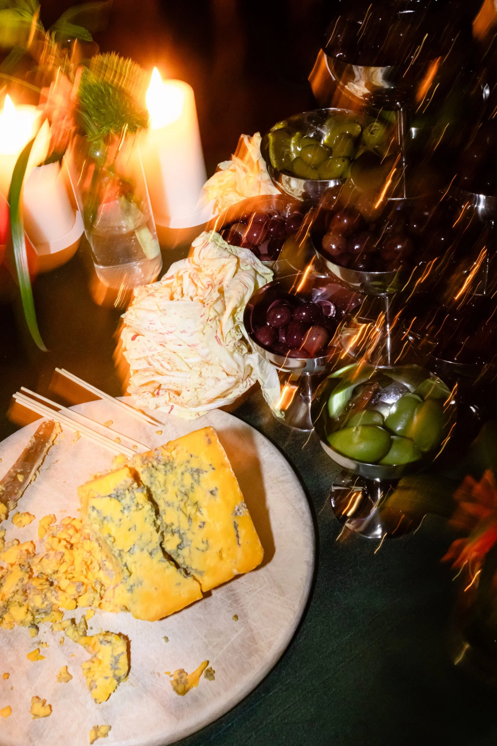 A cheese wheel on a plate, bowls of green and purple grapes, a glass of water with a flower, lit candles, and some sauerkraut or shredded vegetables on a table.