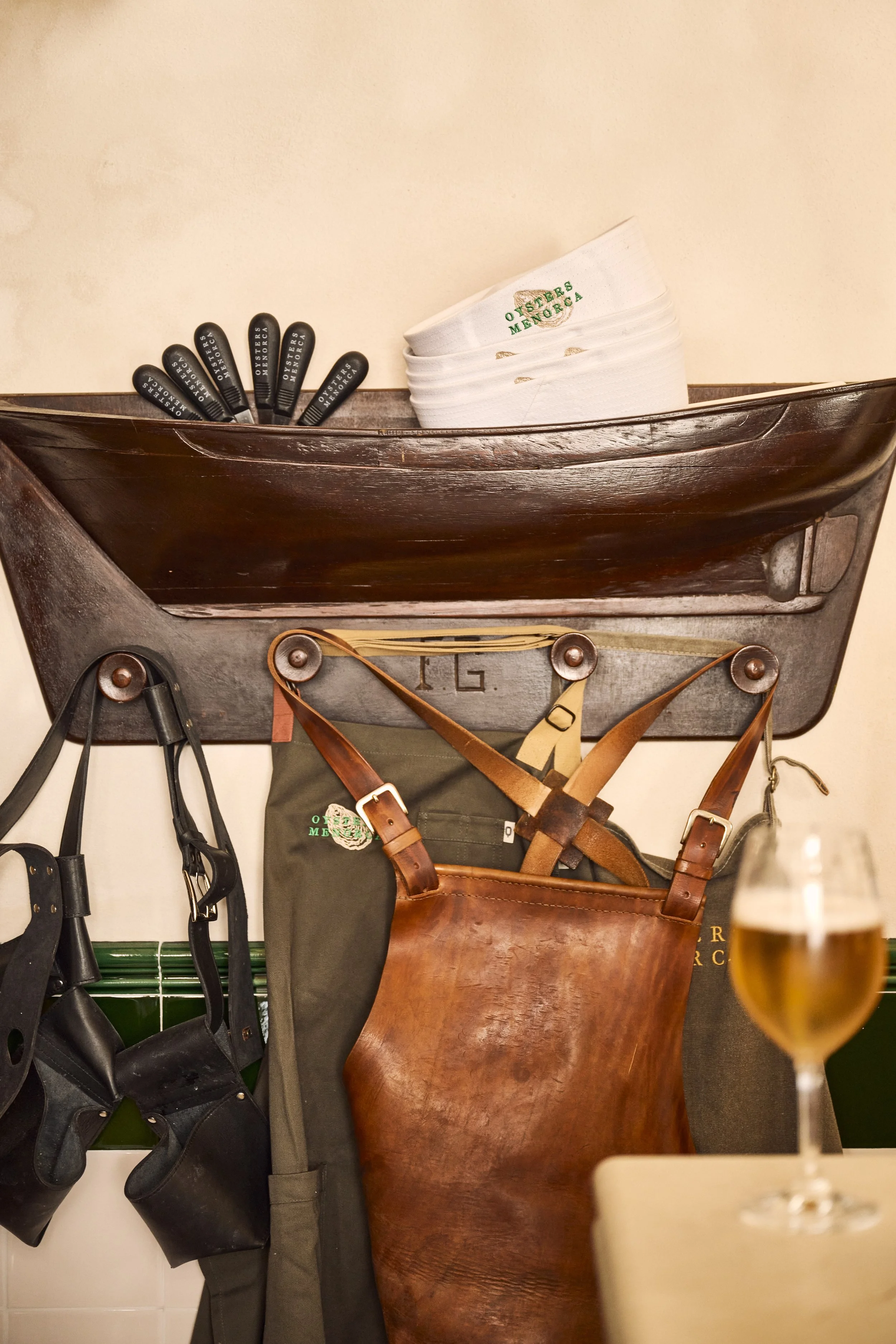 A wall-mounted wooden boat with black and white oyster shucking tools and a stack of oyster shells with a green and white label, along with hanging aprons and leather straps, and a glass of beer in the foreground.
