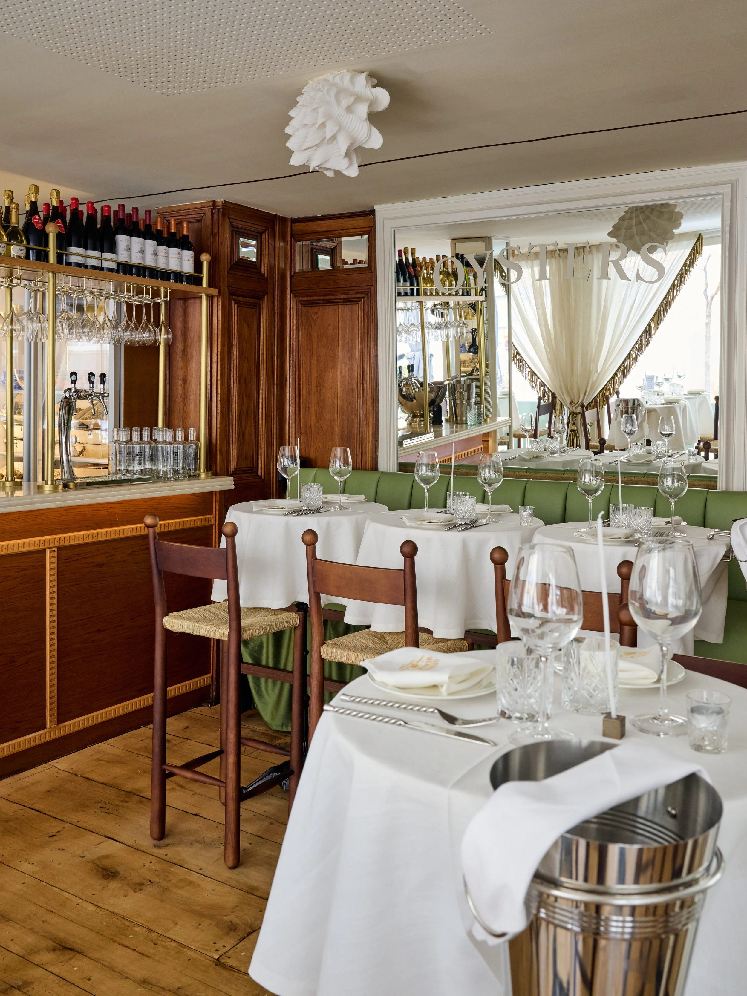 Elegant restaurant interior with tables set with white tablecloths, wine glasses, and silverware, wooden chairs, and a mirror reflecting a bar area with wine bottles and glassware, with curtains and a sign saying 'OYSTERS' in the background.