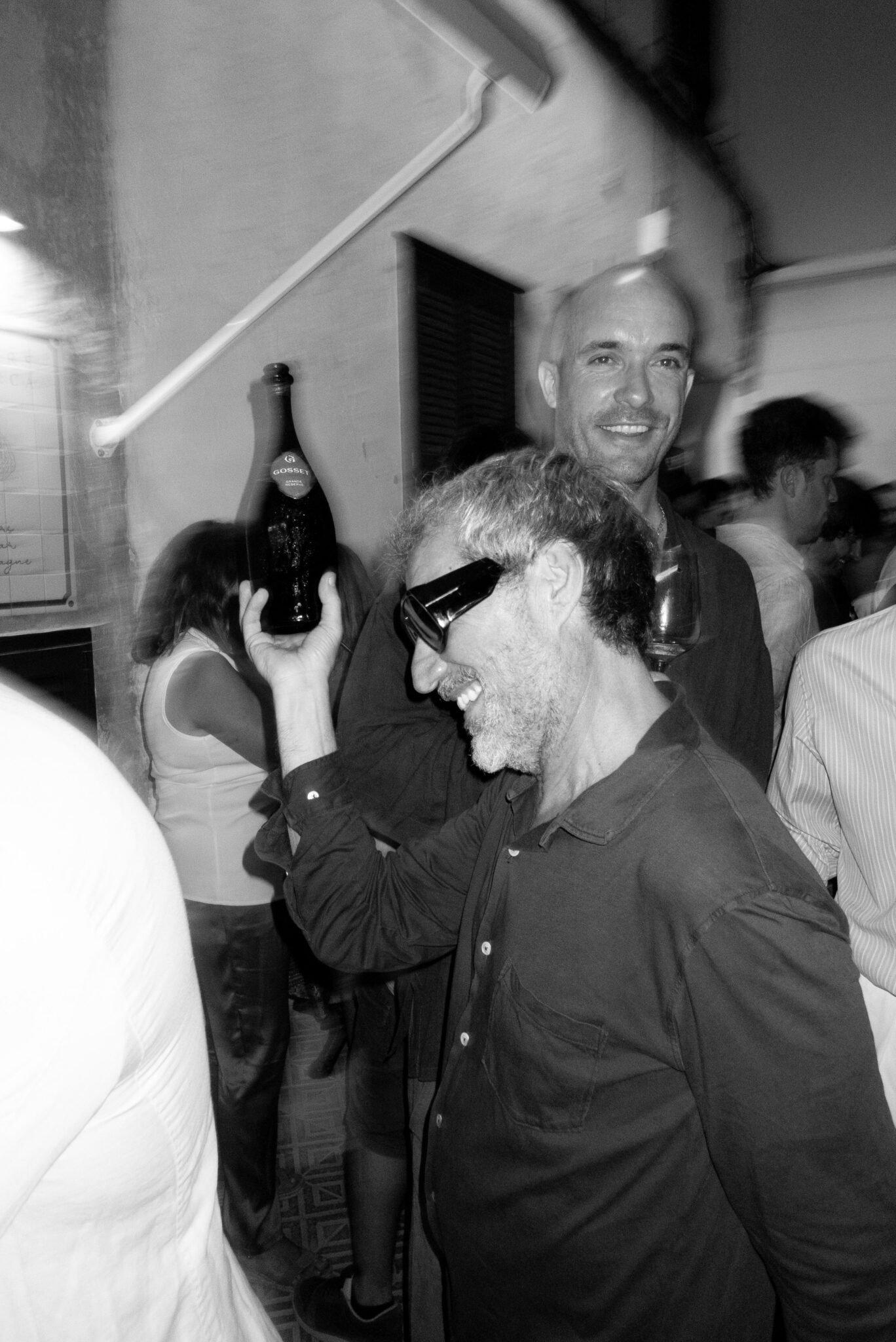 Two men at a lively party, one smiling with a glass of wine, the other holding a bottle of Gosselin beer on his shoulder, surrounded by guests.