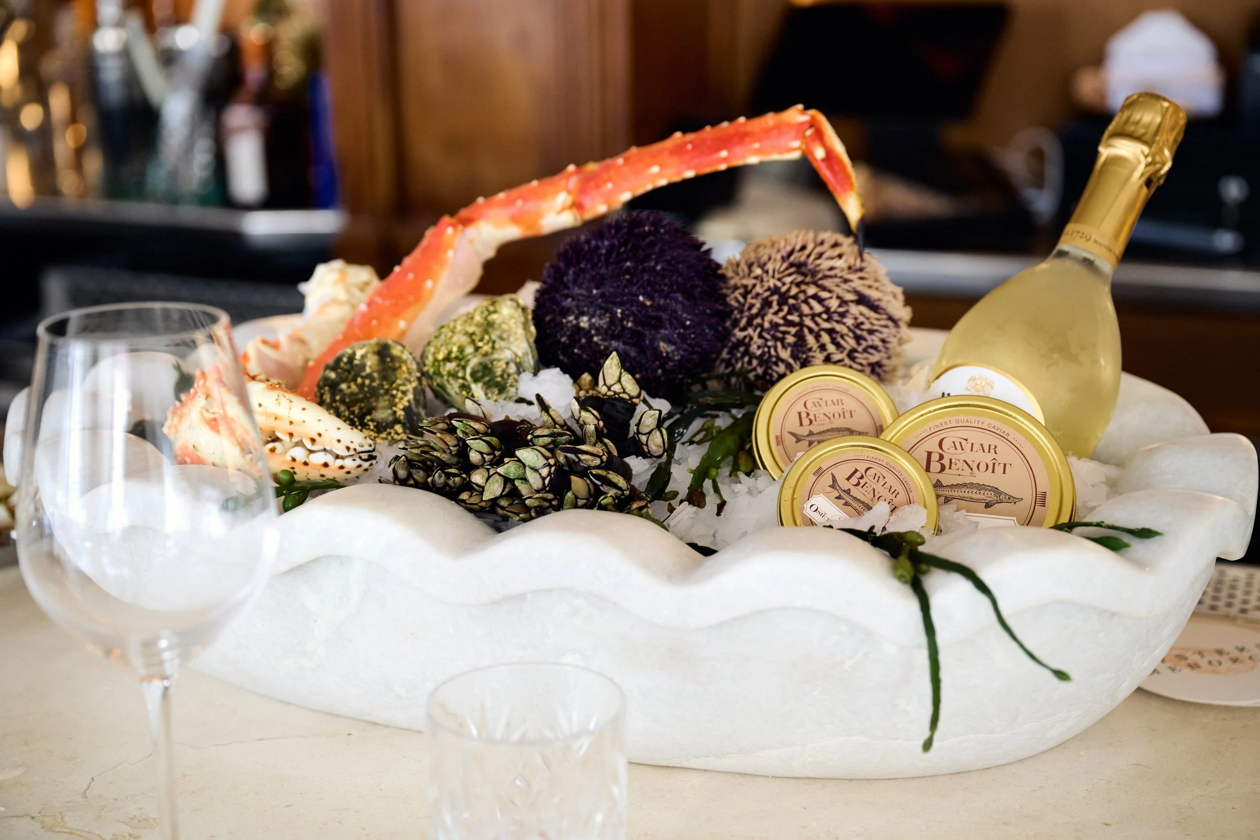 Seafood platter with crab legs, various shellfish, and caviar, served with a bottle of white wine on a bed of ice in a white decorative bowl.
