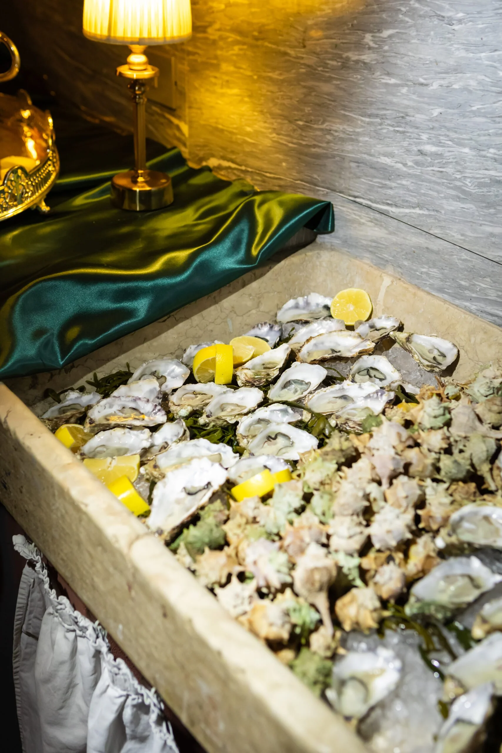 A tray of opened oysters with lemon wedges on ice, next to a decorative cloth and a lamp.