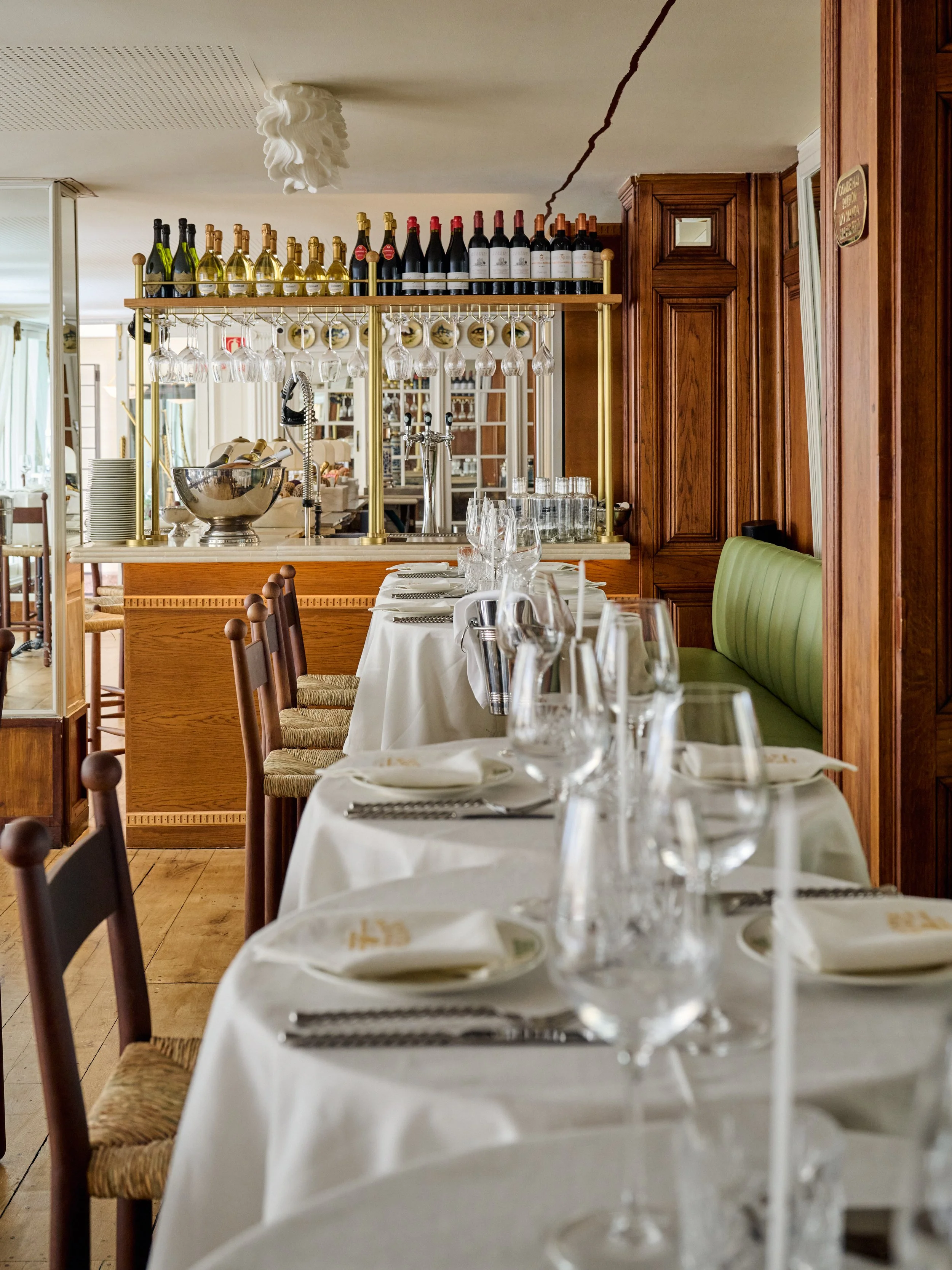 Elegantly set dining table with white tablecloth, wine glasses, and silverware in a restaurant with wooden chairs and a wine display and bar in the background.