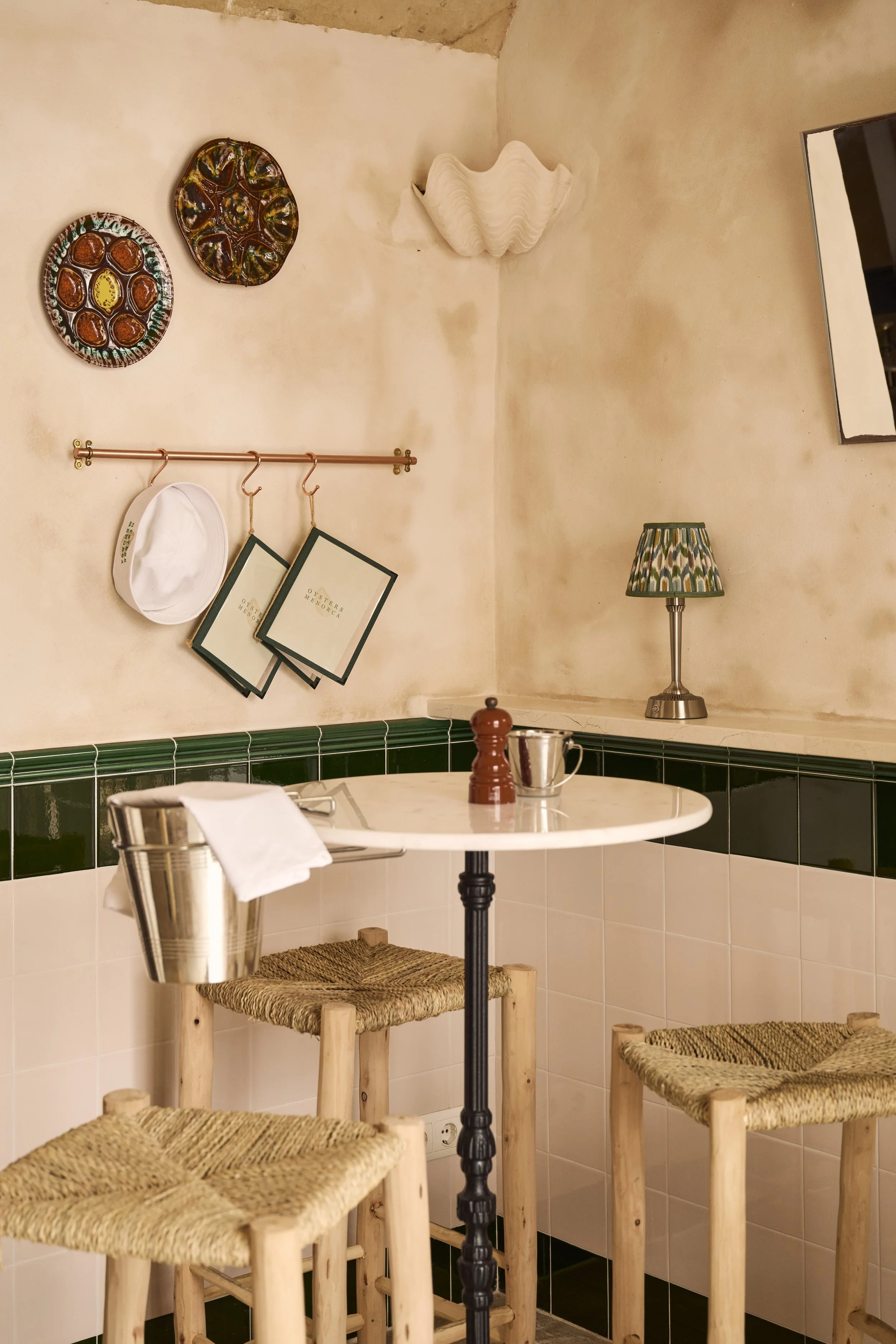 A cozy Italian-style cafe corner with a small round marble-topped table, three woven straw stools, a wall-mounted copper rod with a white hat and menus, decorative plates, a wall sconce, a lamp with a patterned shade, and a mirror.