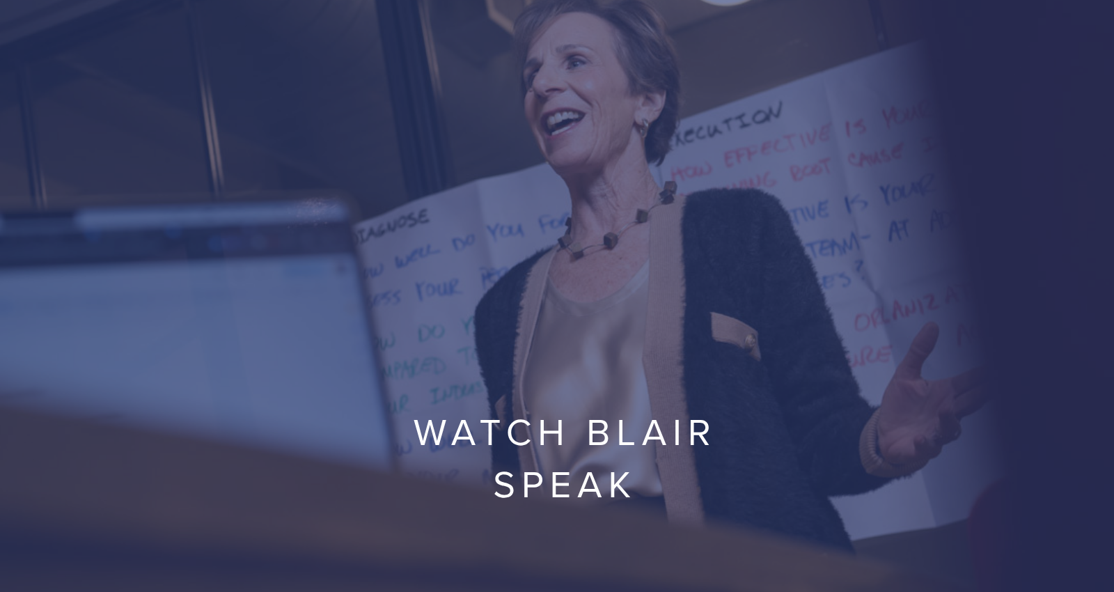 A woman standing in front of a whiteboard, speaking and gesturing with her hand, during a presentation or lecture.
