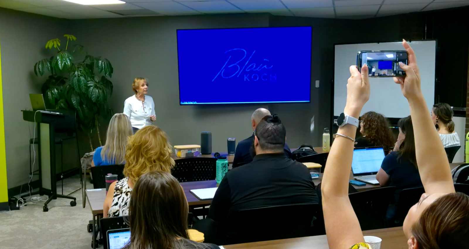 A woman giving a presentation in a classroom with a large screen displaying the name Blair Koch, in front of seated attendees, some taking photos or using laptops.