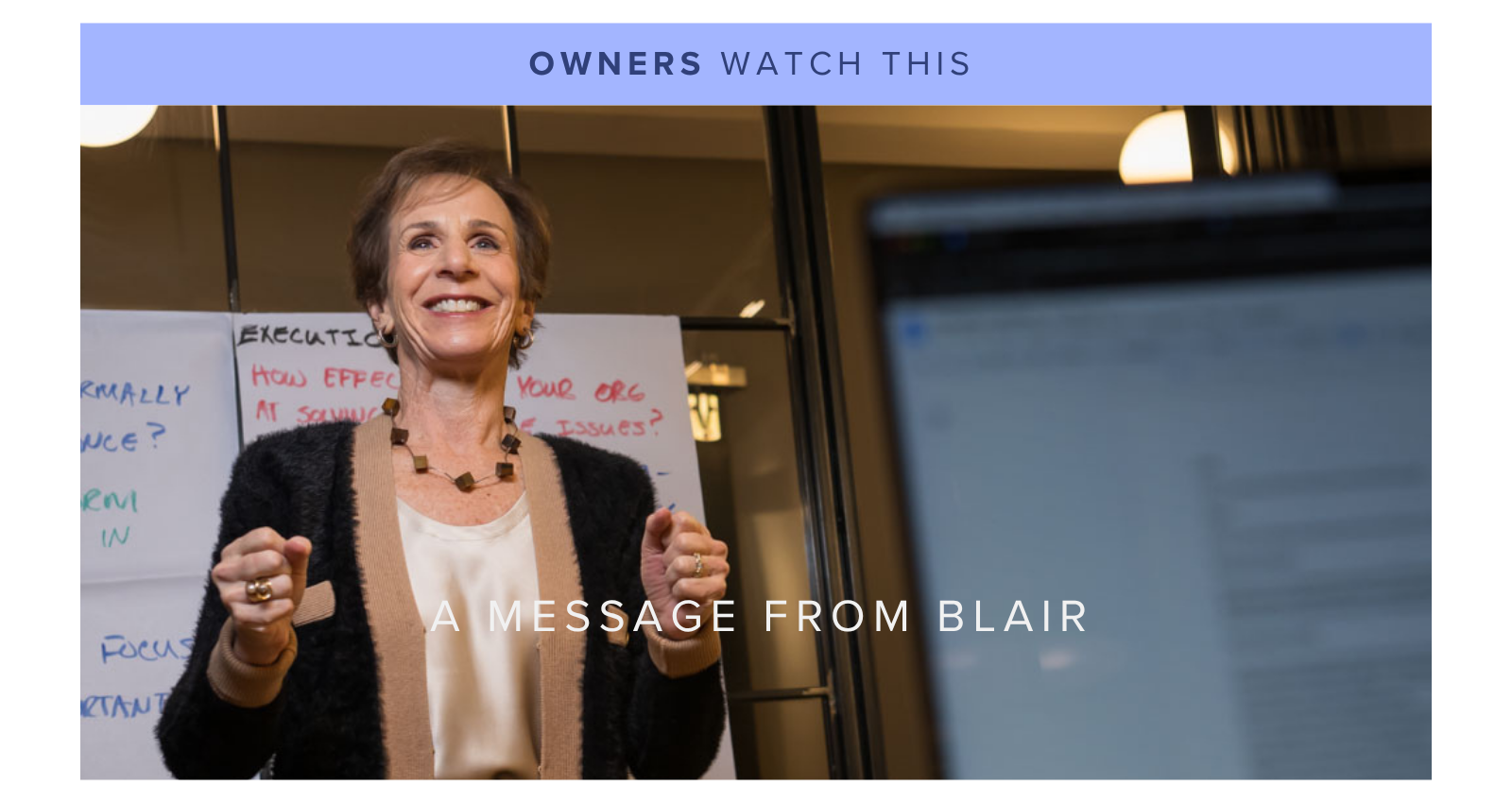 Photo of a smiling woman giving a presentation in an office setting, with a whiteboard behind her containing handwritten notes, and a blurred computer screen in the foreground.