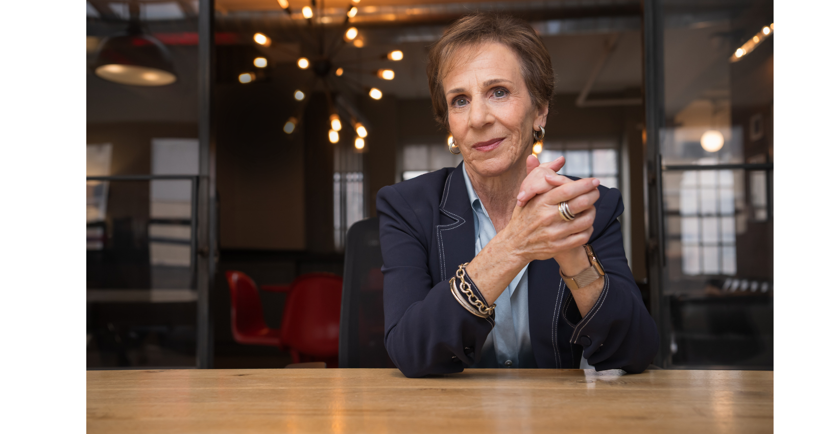 A mature woman with short brown hair sitting at a wooden table in a modern office or cafe environment. She is wearing a navy blazer, light blue shirt, and jewelry. She looks confident and attentive, with her hands clasped together in front of her. The background features warm lighting, glass walls, and some red chairs.