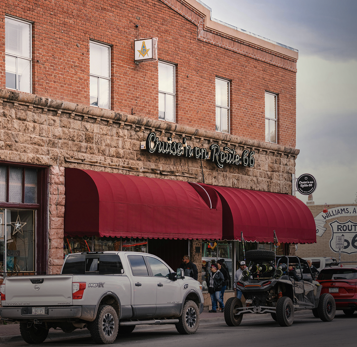 People standing on the sidewalk in front of a building with a sign that reads 'Cruise on Route 66,' red awnings, parked trucks and cars, and a large mural of a Route 66 sign on a neighboring building.