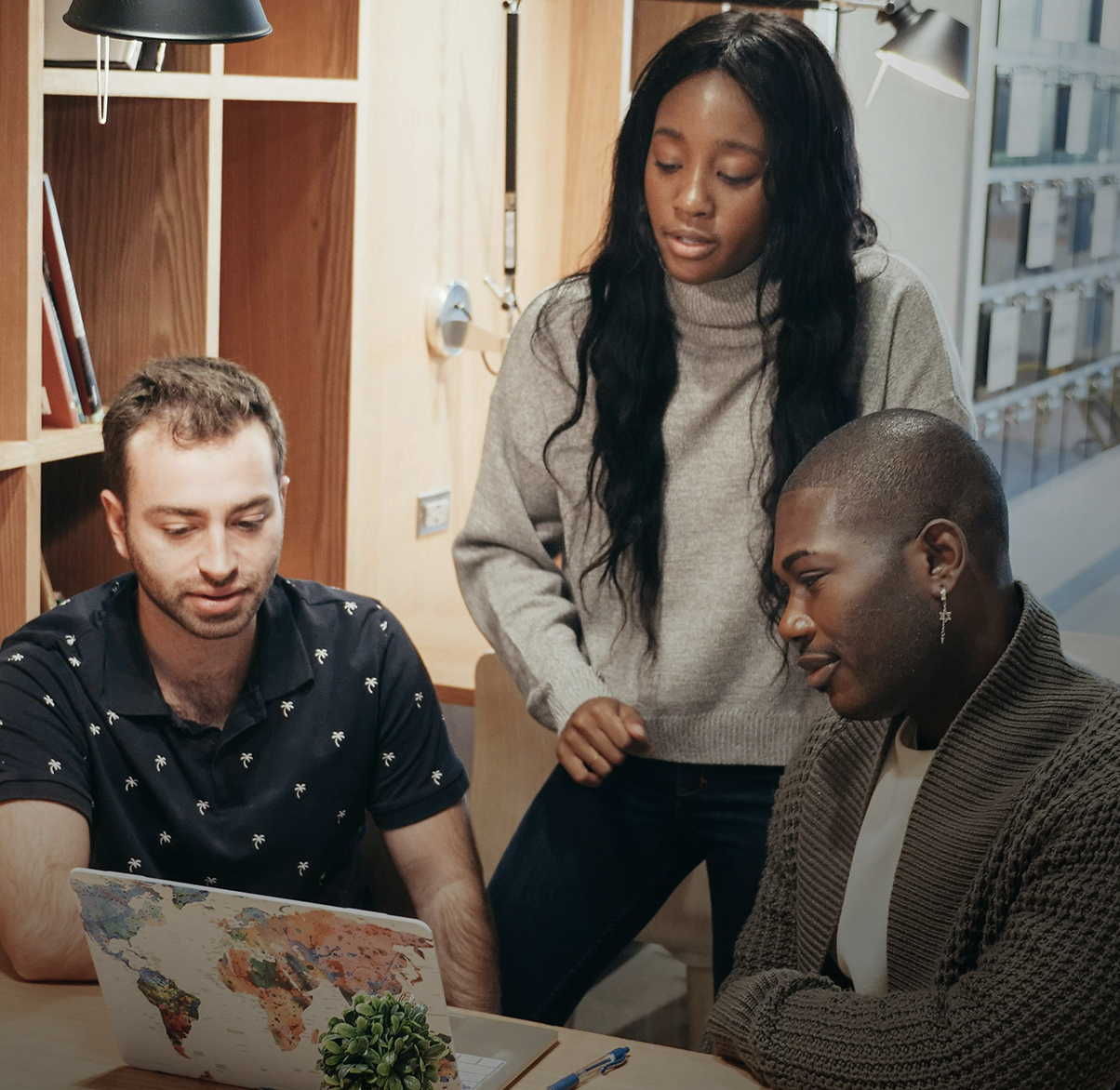 Three young adults working together on a laptop with a world map design, in a modern office or library setting, engaging in a collaborative activity.