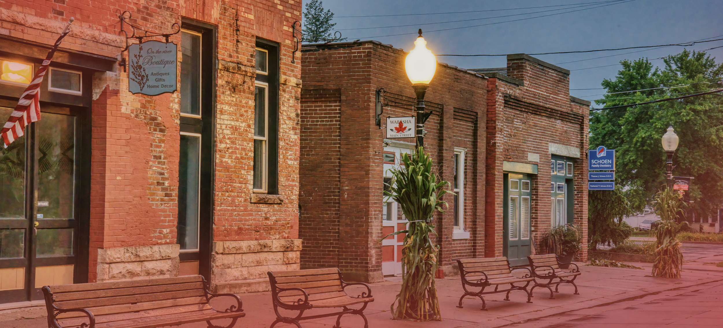 A small town street scene during early evening with brick buildings, benches, street lamps, and decorative corn stalks. Signs for a boutique, a dental office, and a restaurant are visible.