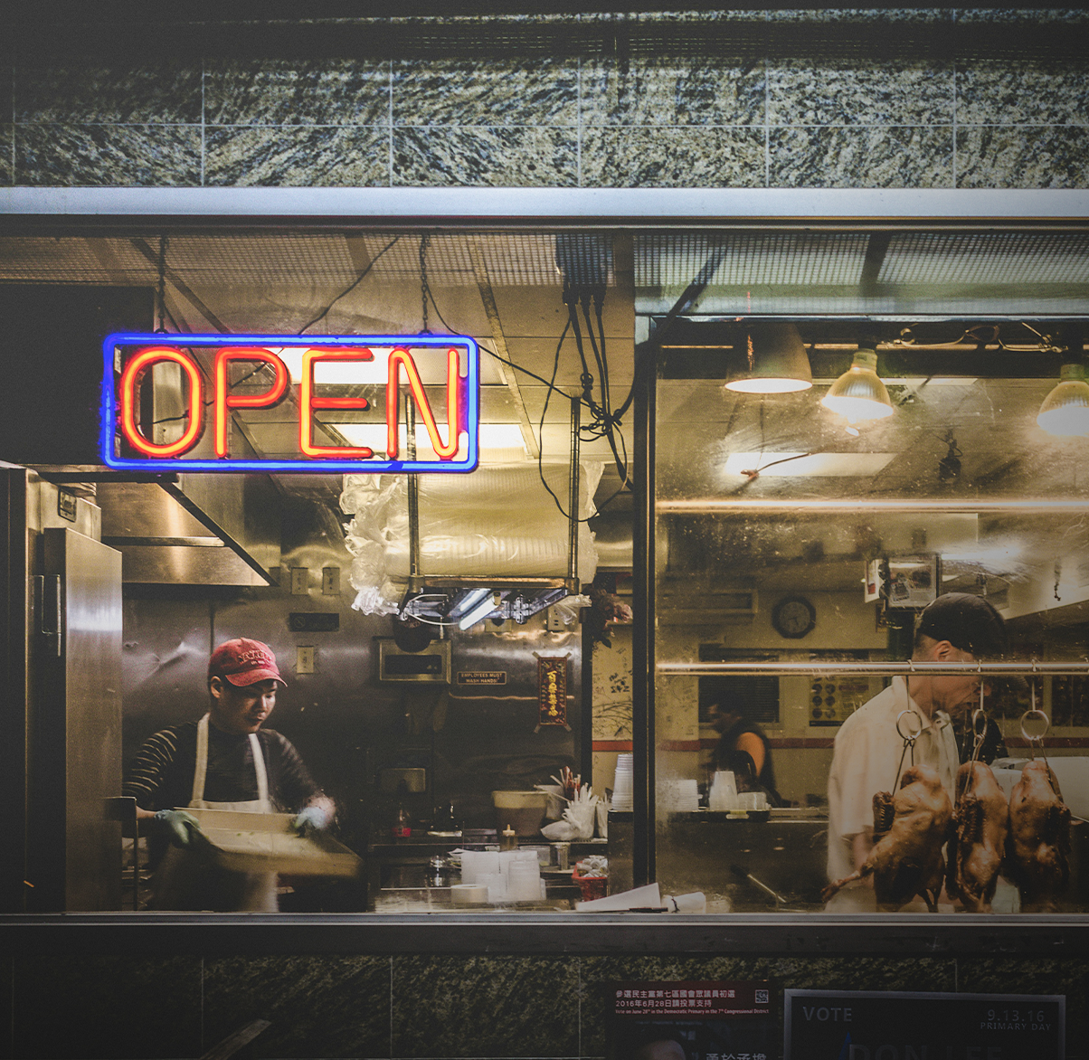 View of a restaurant kitchen through a glass window with an illuminated red and blue 'OPEN' sign, showing two staff members preparing food; one is cooking and the other is handling hanging roasted chickens.