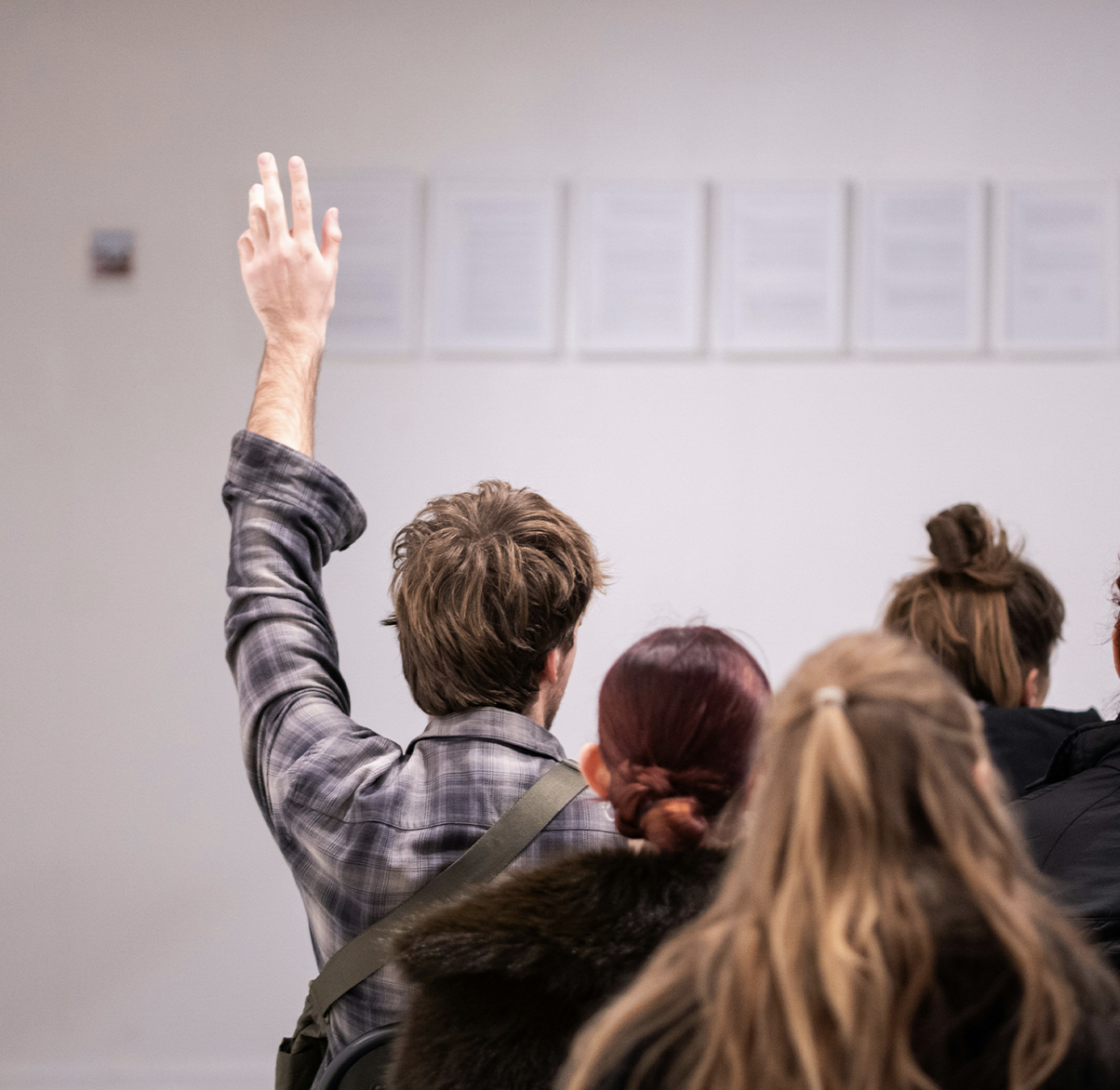 A person with brown hair and a plaid shirt raising their hand in a classroom setting surrounded by other students.