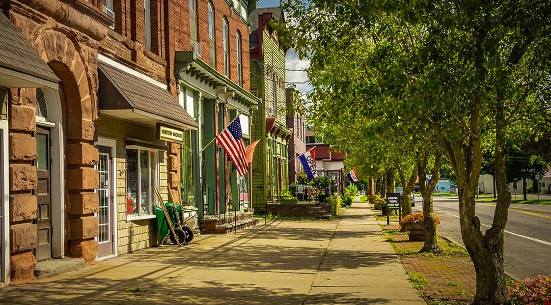 Downtown street with historic brick buildings, American flags, and trees lining the sidewalk.
